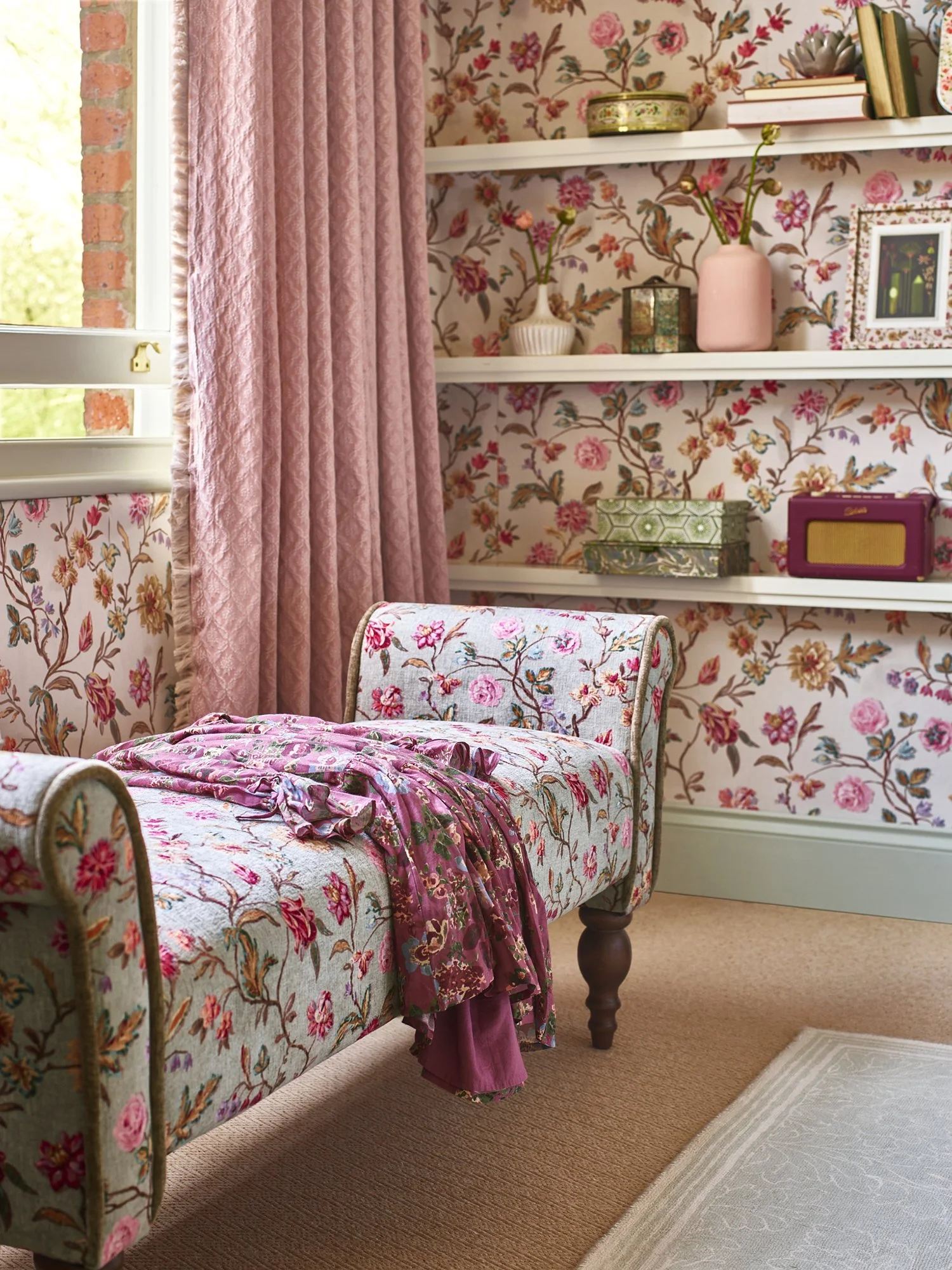 A cozy bedroom with floral wallpaper, pink curtains, a vintage floral upholstered chaise lounge with a pink throw, white shelves with decorative boxes, vases, and framed photos near a window with natural light.