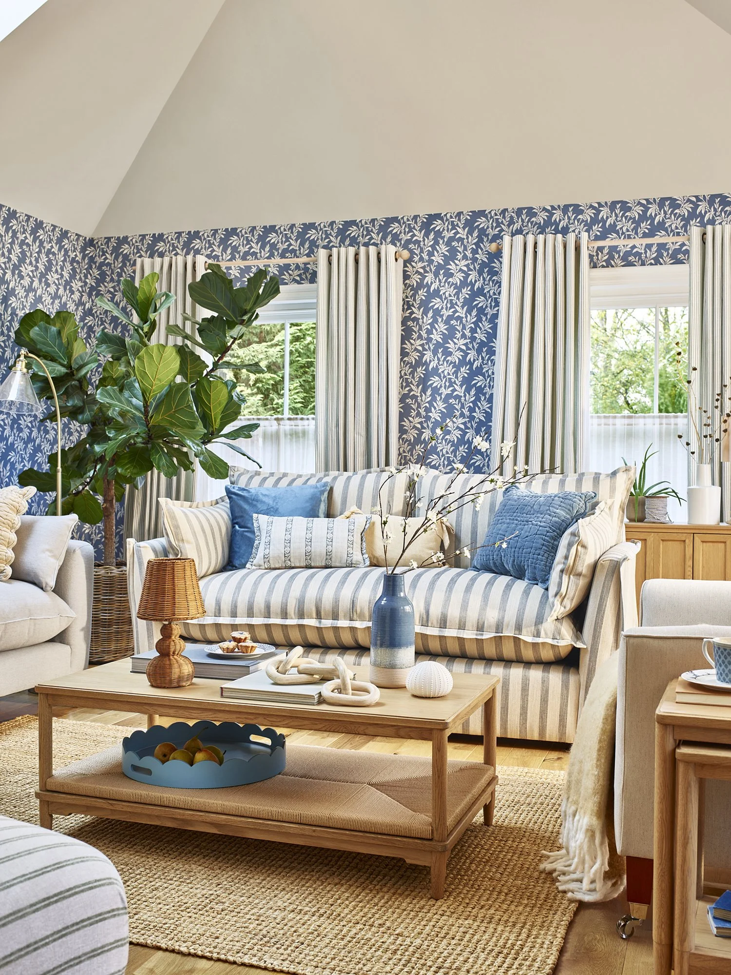 Living room with blue floral wallpaper, striped curtains, a striped sofa with blue pillows, a wooden coffee table with decorative items, a large green potted plant, and a window showing greenery outside.