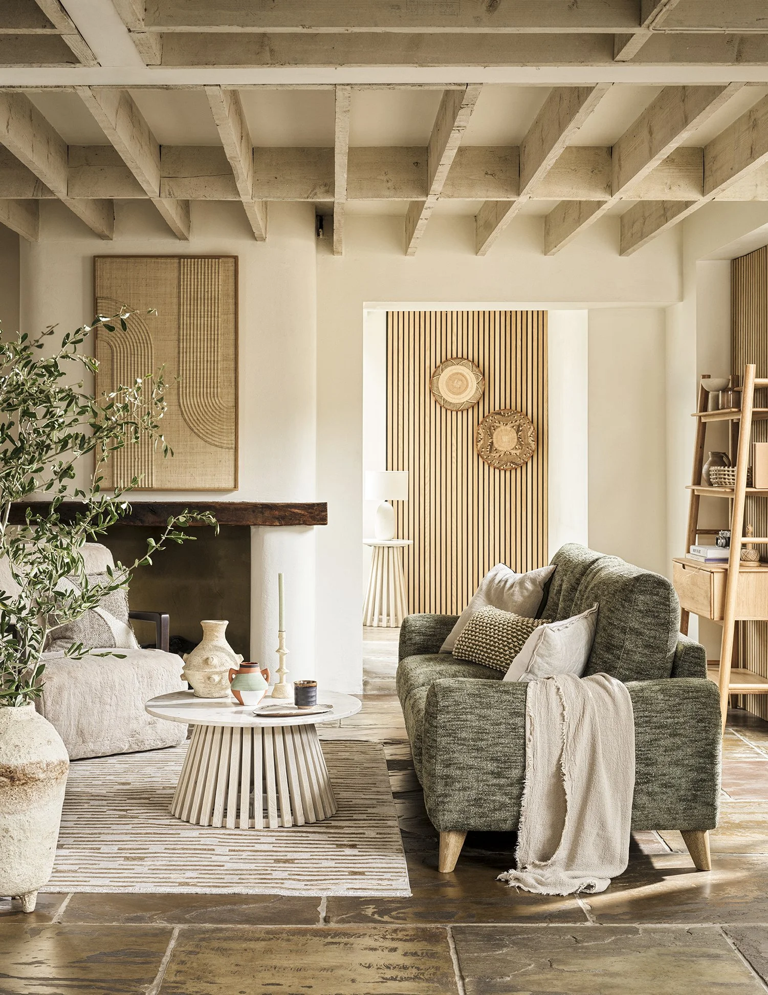 Living room with dark green sofa, white pillows, cream throw blanket, wood floor, beige rug, marble-topped coffee table, fireplace with wooden mantel, wall art, houseplants, wooden shelves, and decorative wall hangings.