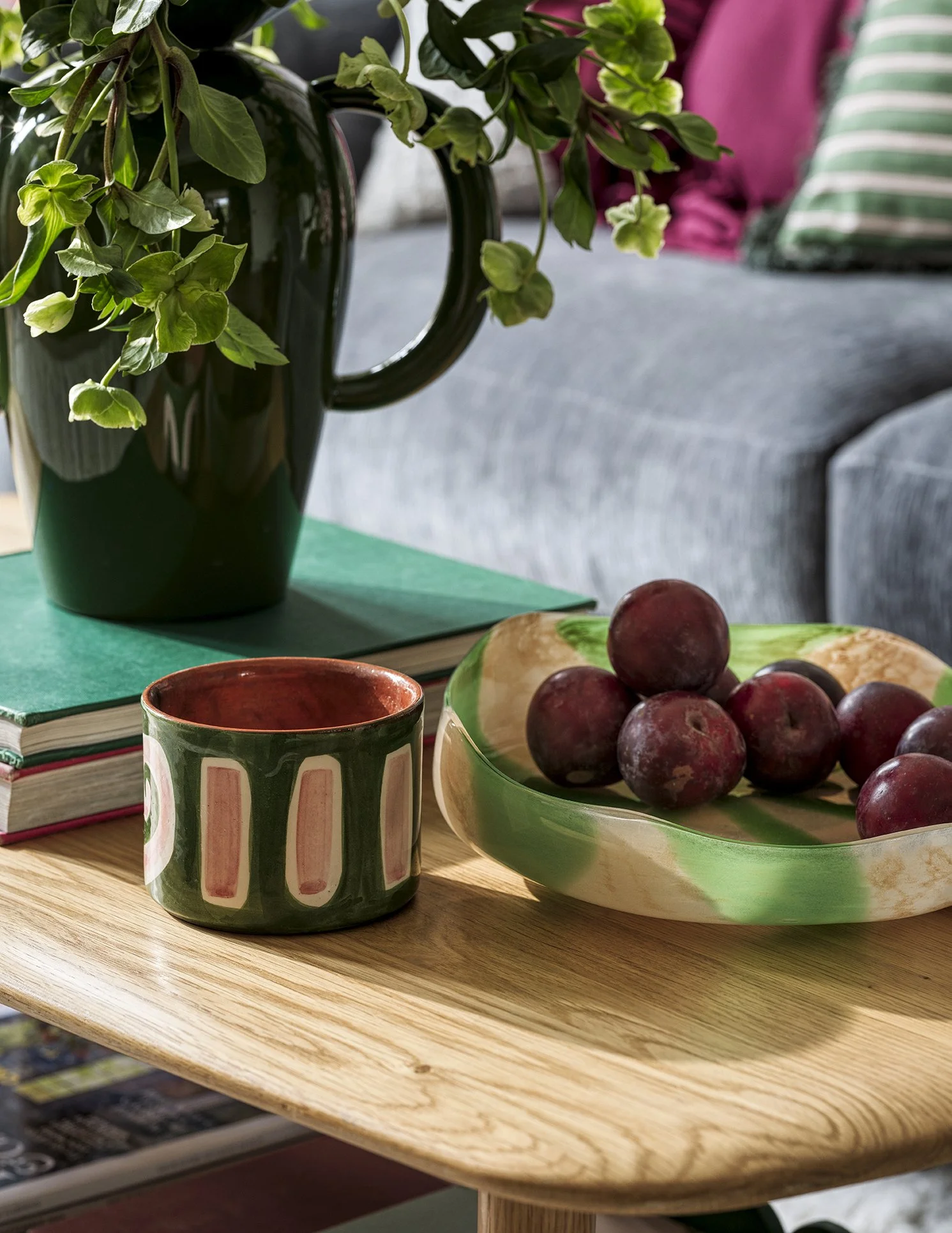 A green ceramic mug, a glass bowl filled with plums, a black vase with green leafy plants, and a stack of books on a wooden table, with a gray sofa and pink and green pillows in the background.