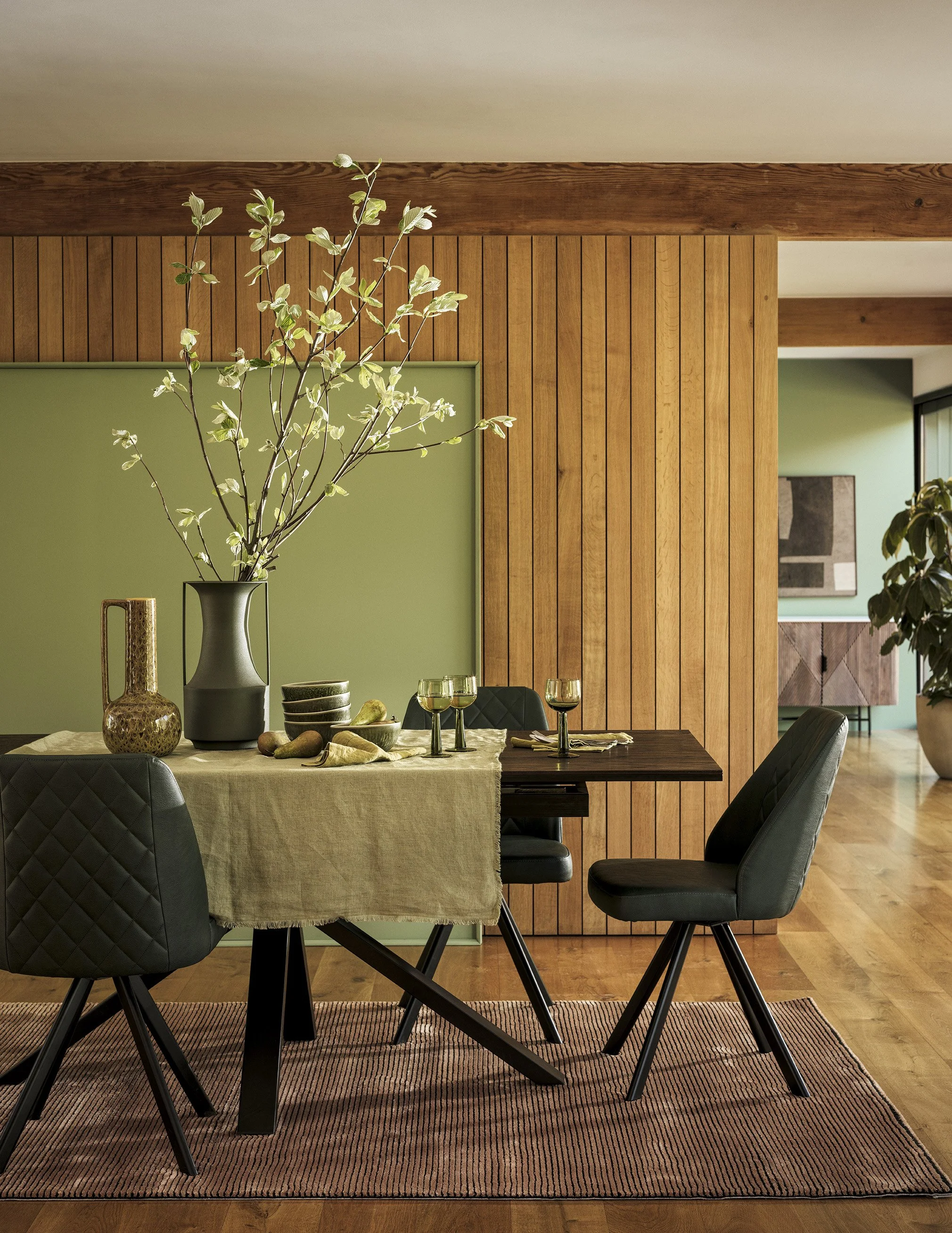 Modern dining room with black chairs, a wooden table, a beige tablecloth, a large vase with greenery, and a green wall with wood paneling.