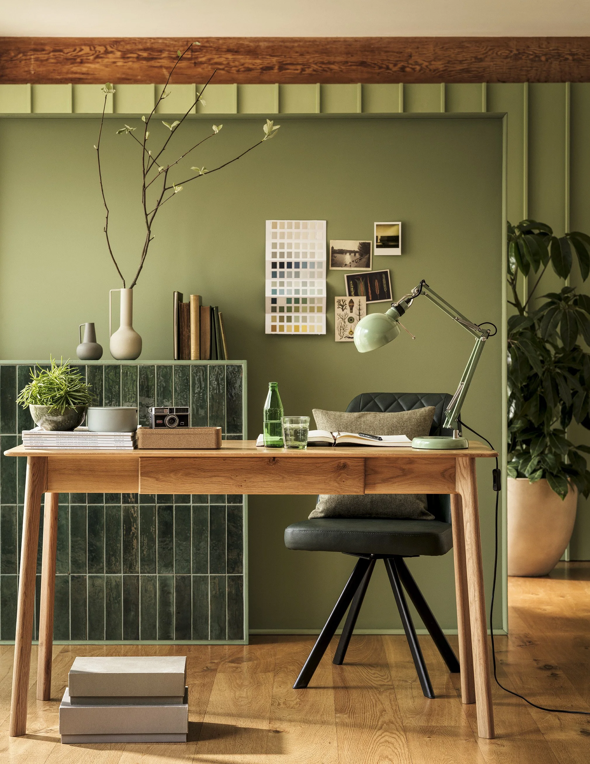 A home office with a wooden desk, black chair, green wall, desk lamp, books, plants, photographs, color swatches, and a large potted plant in the corner.