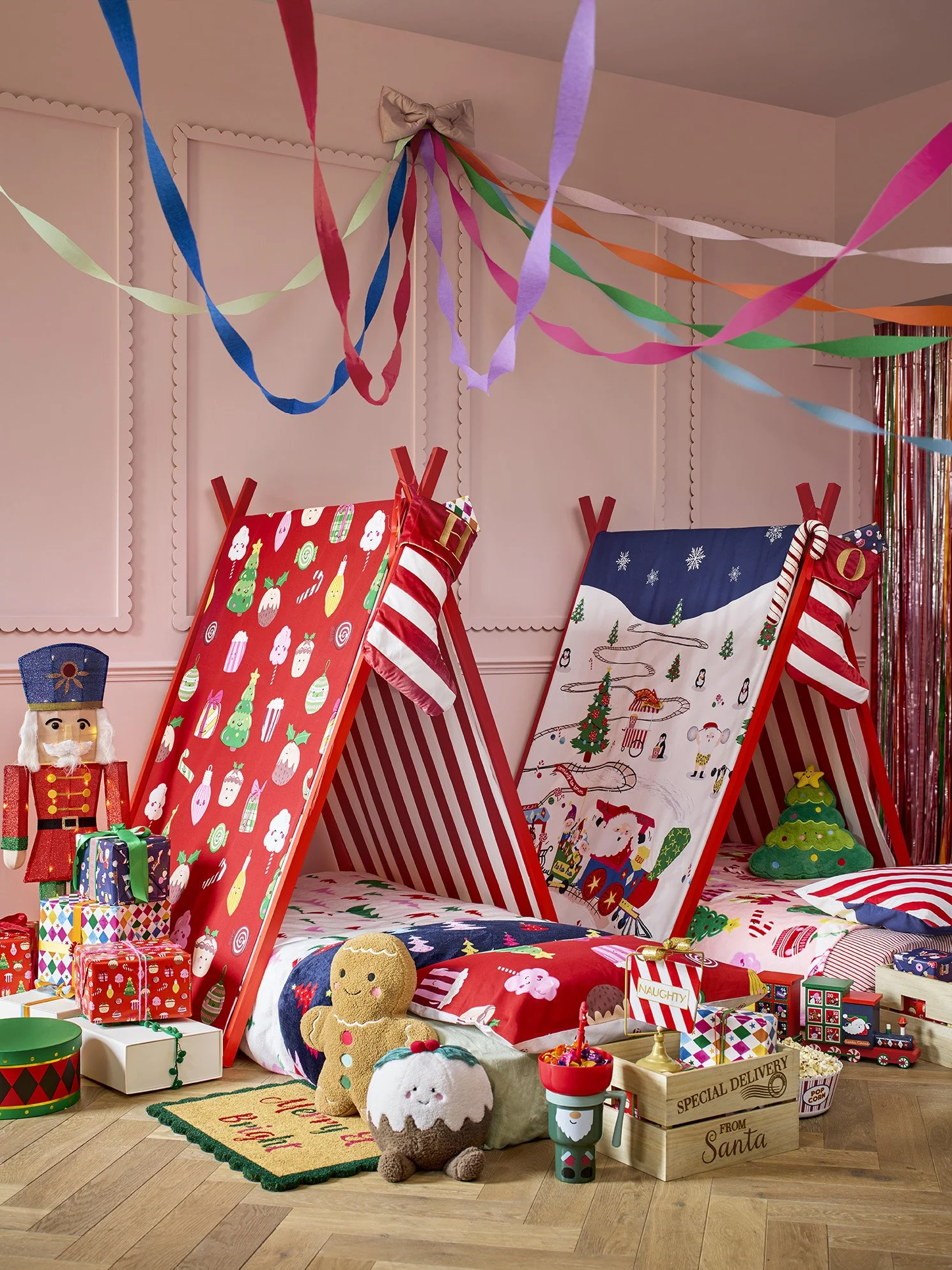 Christmas-themed bedroom decorated with colorful streamers, two beds with striped red and white bedding, Christmas stocking, toy nutcracker, presents, plush gingerbread, cupcake, Christmas tree pillow, and other holiday decorations.