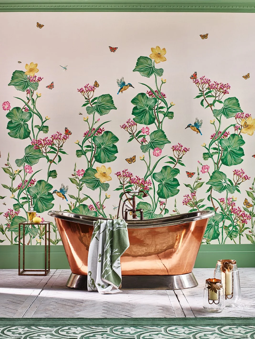A bathroom with a copper bathtub in front of a floral wallpaper featuring green leaves, pink flowers, and yellow blooms, with hummingbirds and butterflies. A small side table holds a drink and a lemon, and two large candles are on the floor.