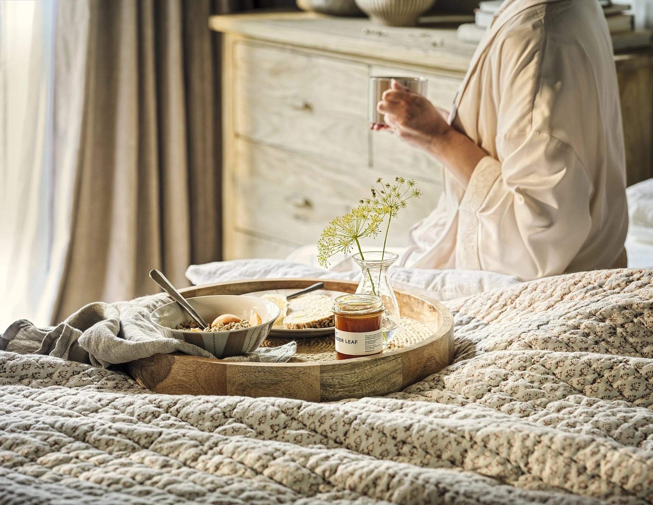 A person sitting on a bed holding a glass of tea or coffee with a breakfast tray in front, containing cereal, bread, a jar of jam, and a vase with flowers in a cozy, sunlit bedroom.