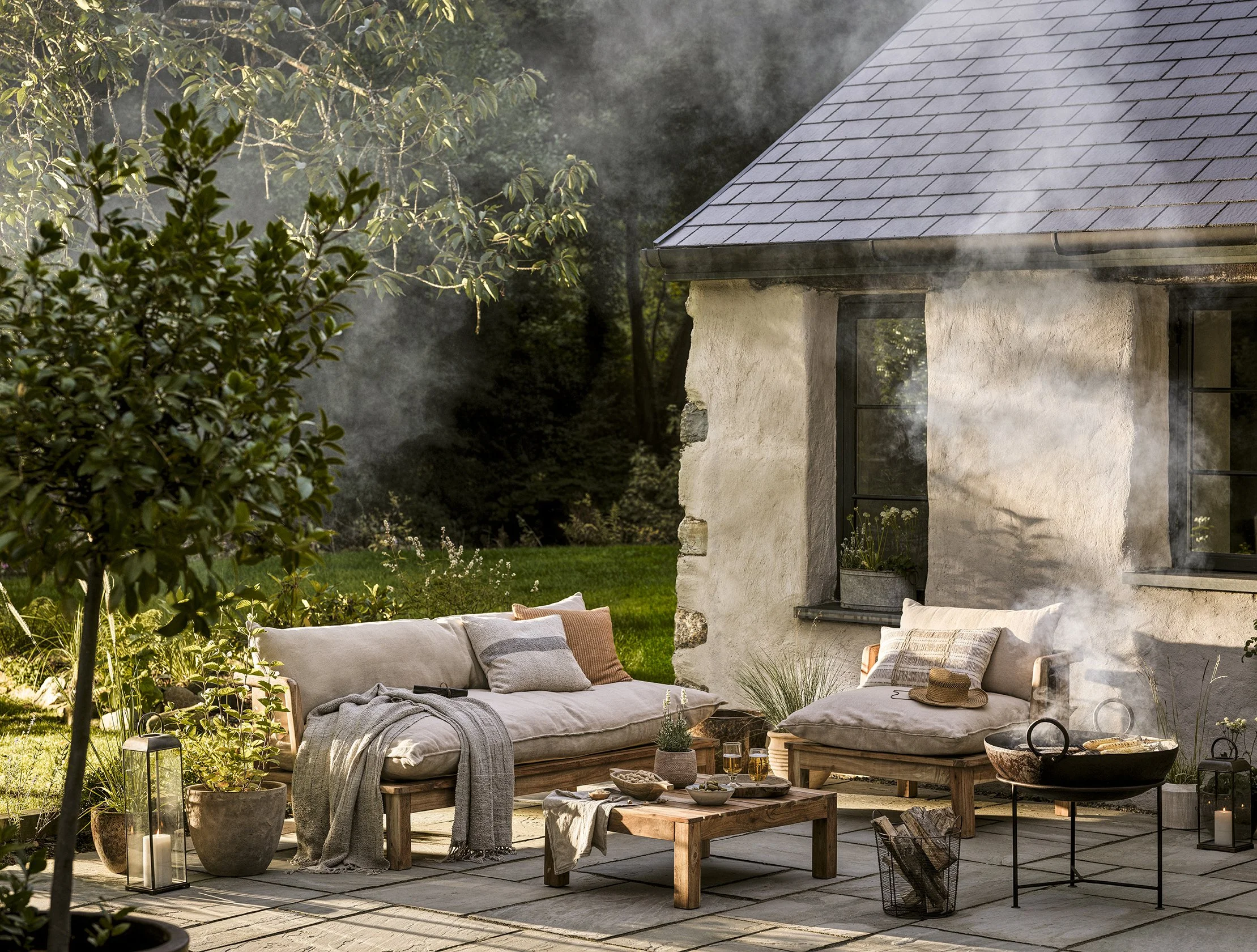 Outdoor patio with beige cushioned sofa and chairs, wooden coffee table, potted plants, lanterns, and candles on stone flooring next to a rustic house with steam rising from the side.