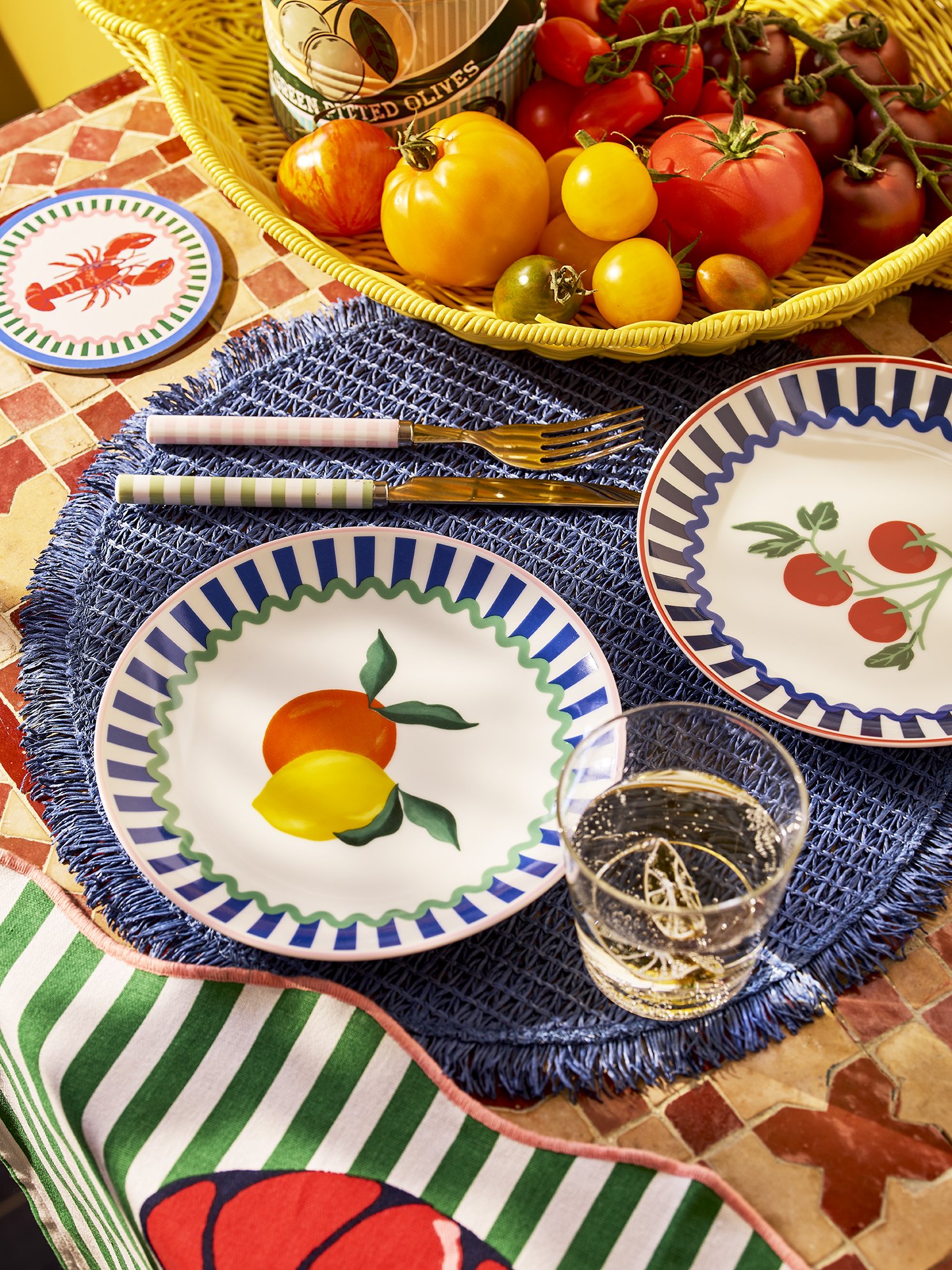 Table setting with colorful fruit-patterned plates, a glass of sparkling water with lemon, and a basket of fresh tomatoes, yellow, red, and green heirloom tomatoes, alongside a can of green salted olives.