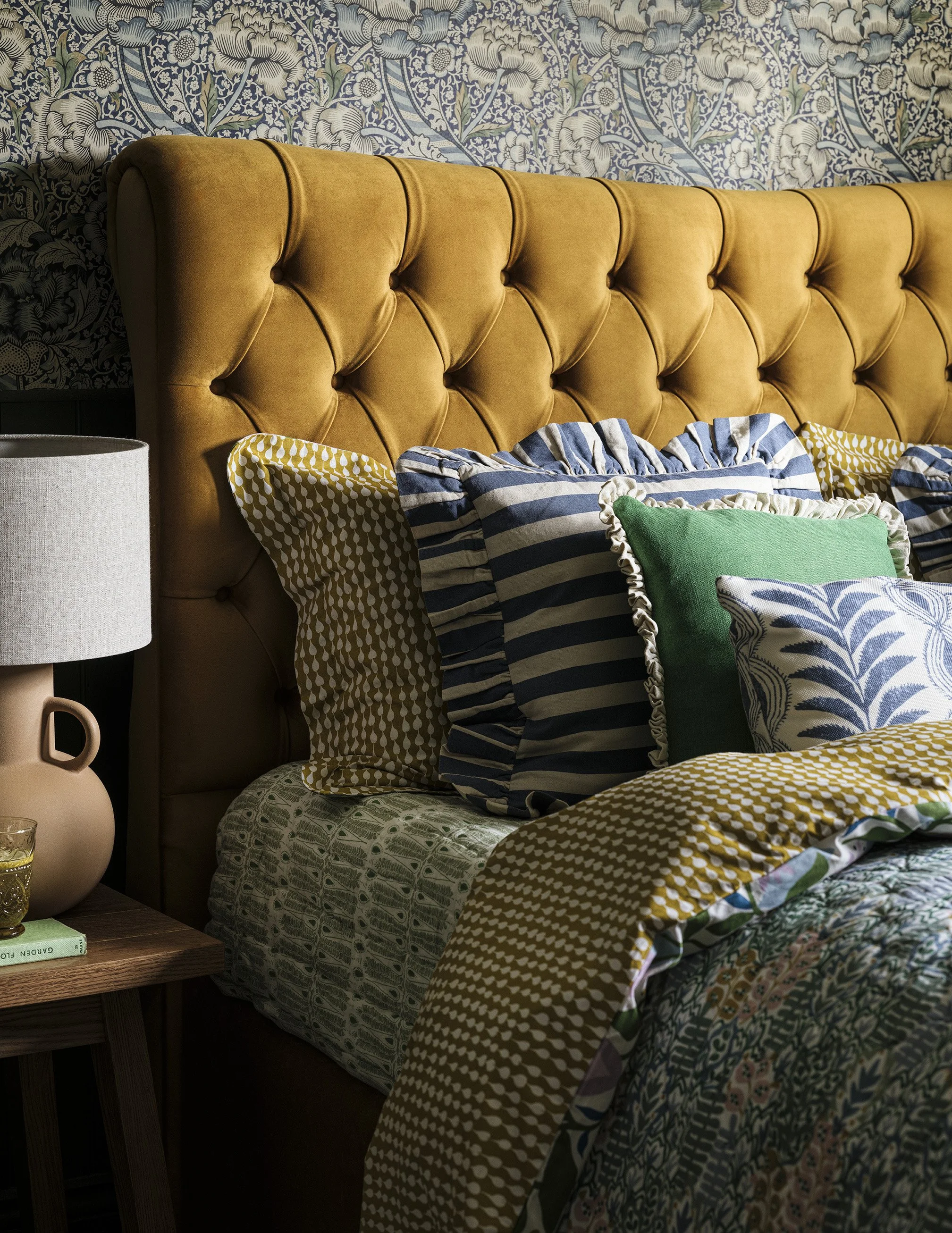 A bedroom with an orange tufted headboard, decorative pillows in green, blue, and yellow patterns, and a bedside table with a beige lamp, glass, and a book, against floral wallpaper.