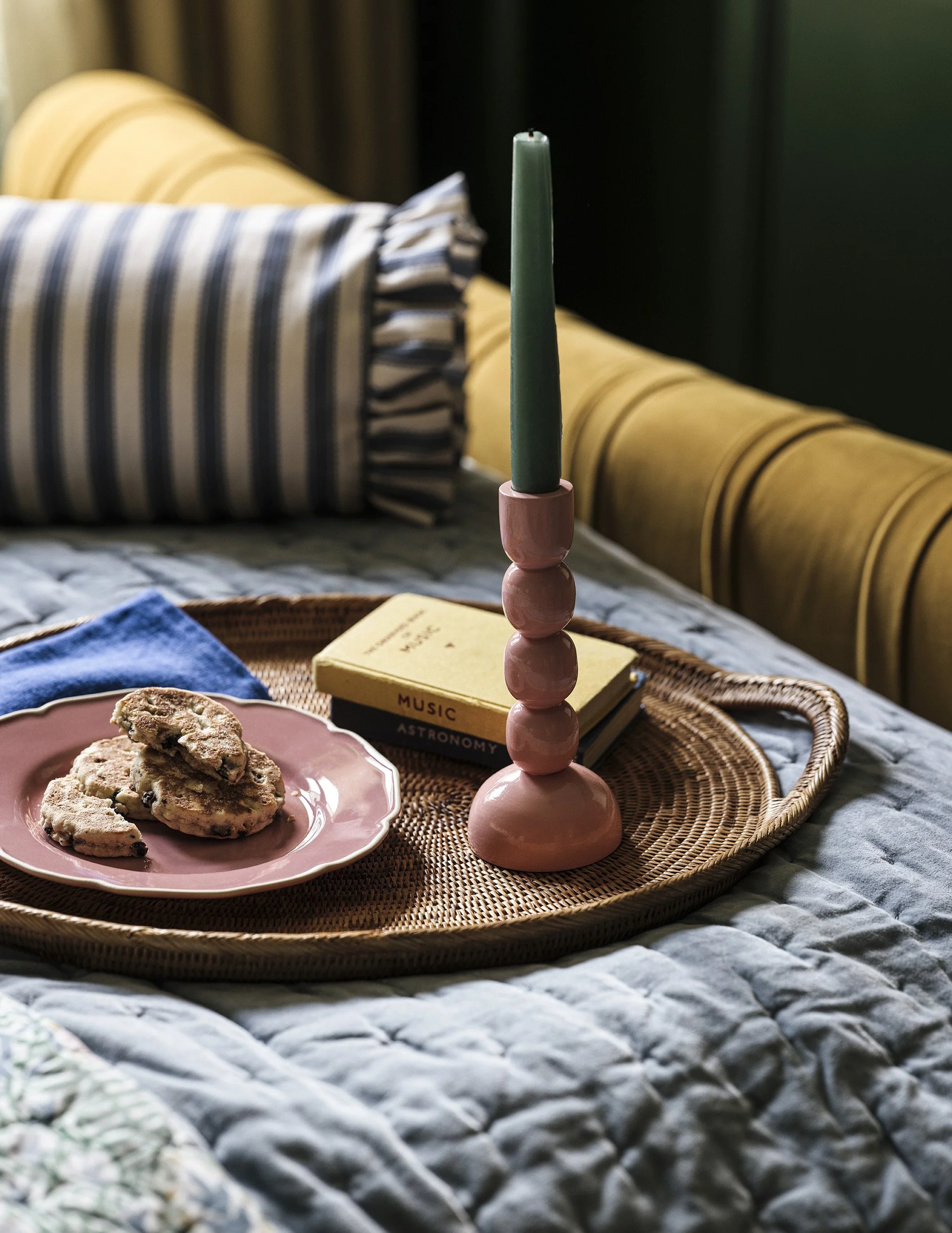 A pink candle holder with a green candle, a pink plate with cookies, a yellow book titled "Music" and "Astronomy," all on a woven tray on a bed with a blue pillow and striped blanket in the background.