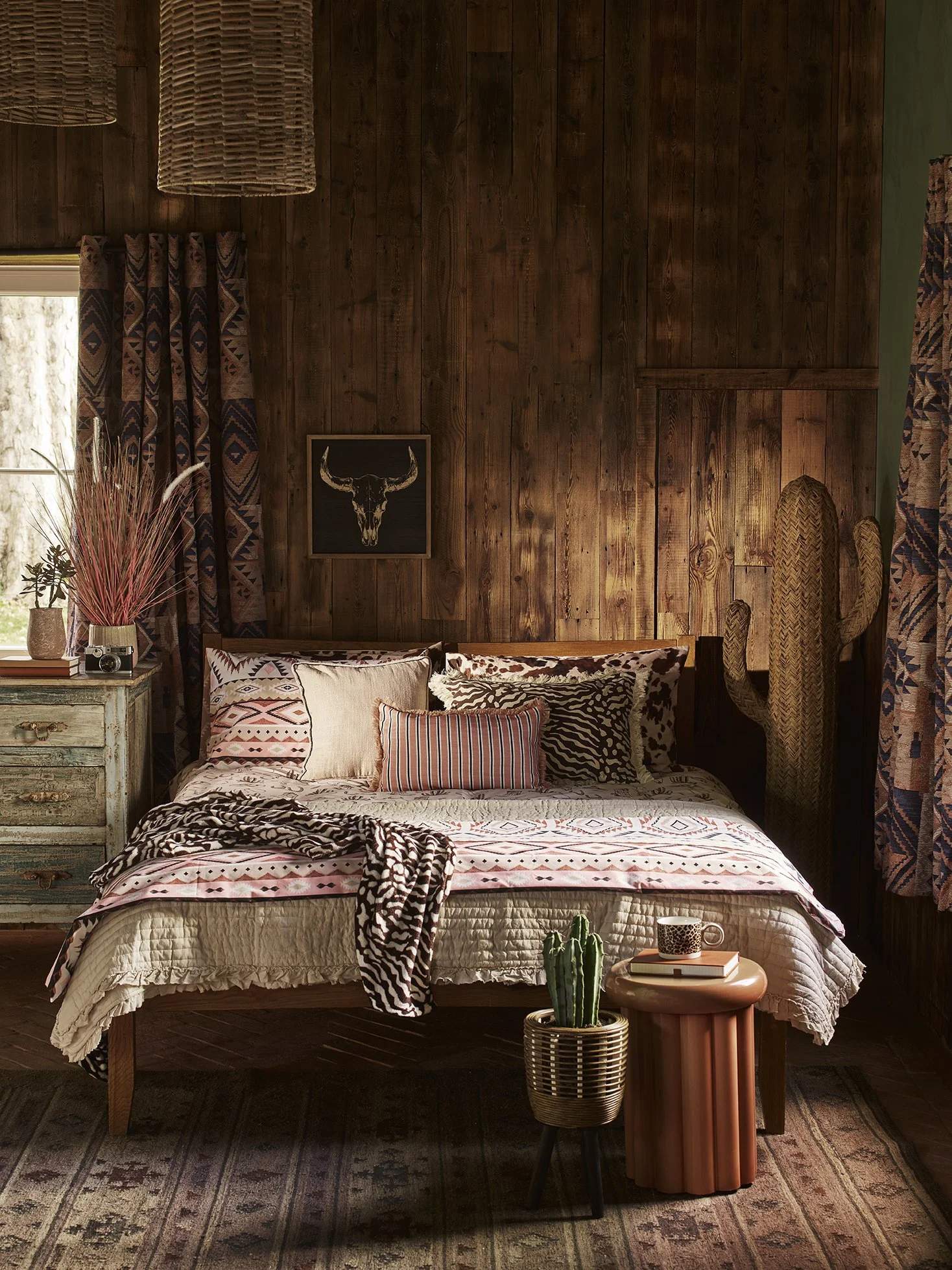 A cozy bedroom with wood-paneled walls, a bed with patterned pillows and a blanket, a green distressed nightstand with plants and a camera, a cactus on a small side table, and decorated curtains and wall art in a rustic style.