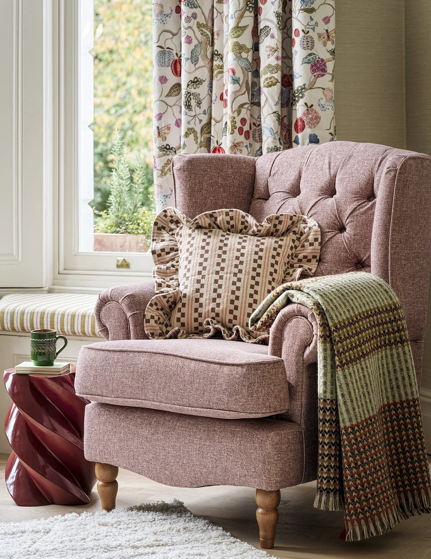 A cozy living room corner with a pink upholstered armchair, decorative pillows, a blanket, a window with patterned curtains, and a side table with a green mug, books, and a sculpture.