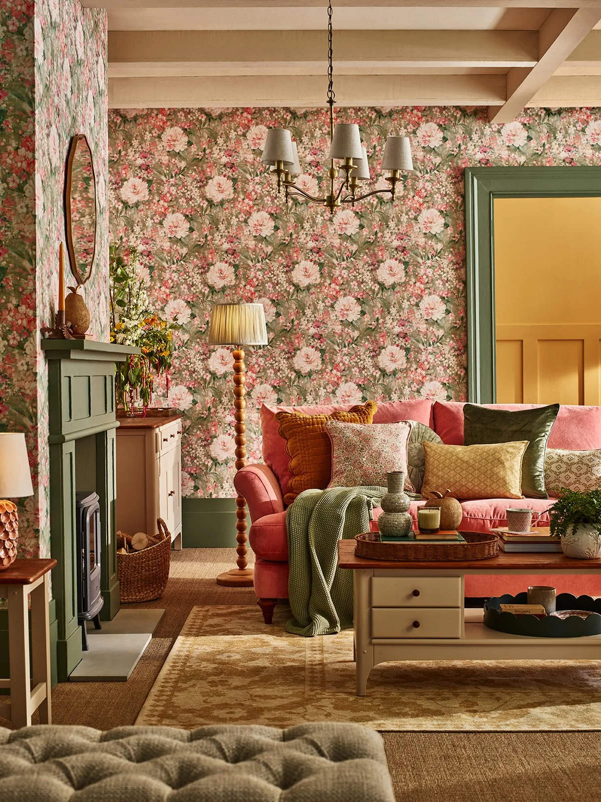 A cozy living room with floral wallpaper, a pink sofa with multiple cushions, a wooden coffee table, a green fireplace, a floor lamp, and a chandelier.