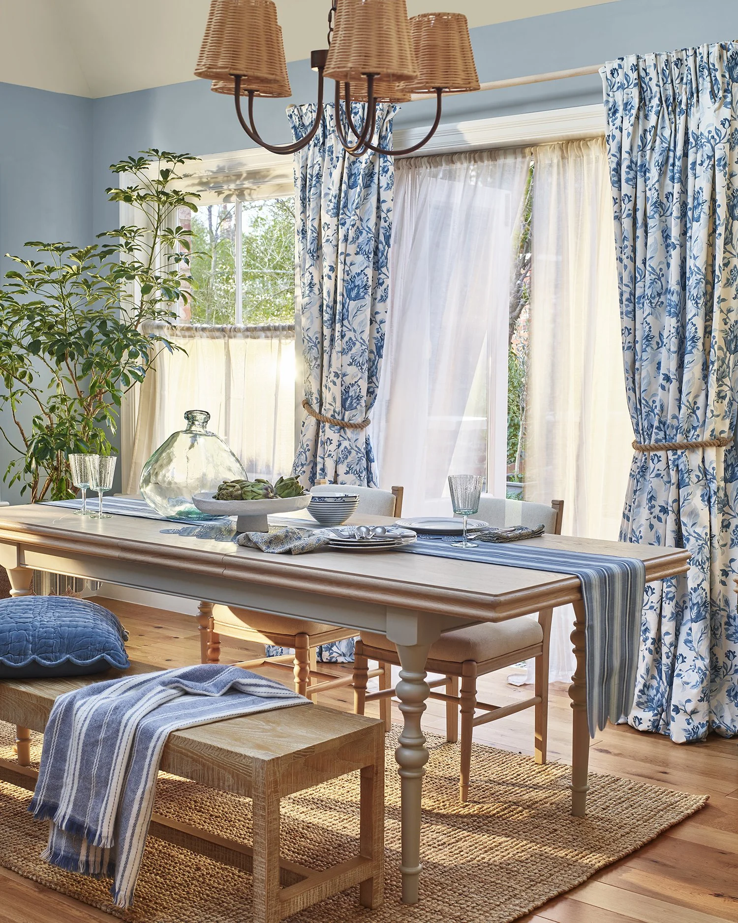 A bright dining room with a wooden table set for a meal, surrounded by chairs. Blue and white floral curtains frame large windows that let in natural light. A large glass jar and serving dishes are on the table, complemented by a striped table runner
