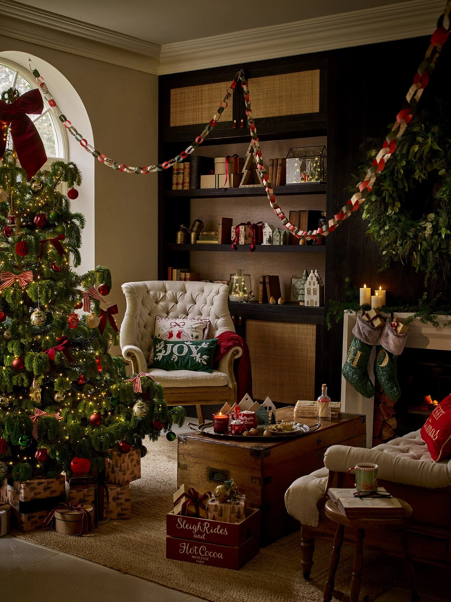 A cozy living room decorated for Christmas with a lit Christmas tree, wrapped presents, stockings hanging on the fireplace, and holiday decorations.