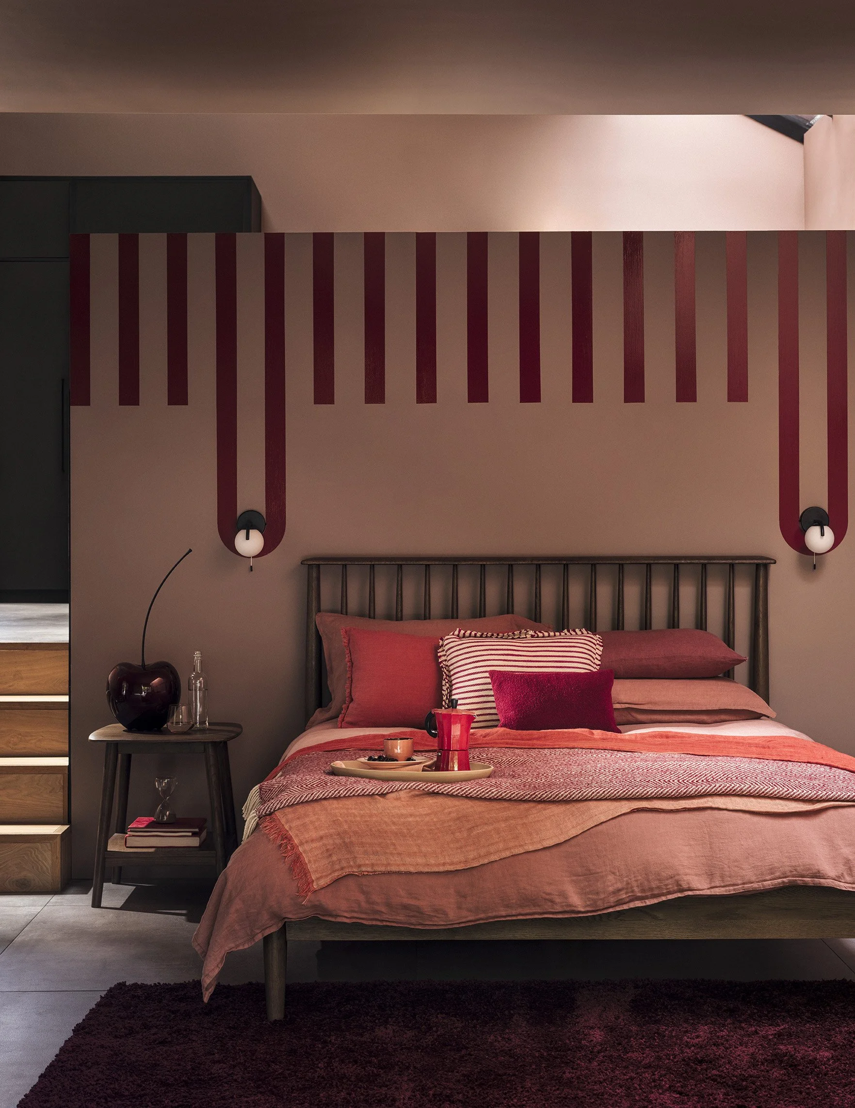 A bedroom with a bed, pillows, and a tray with a drink and snacks. Decor includes red accents and wall lighting.