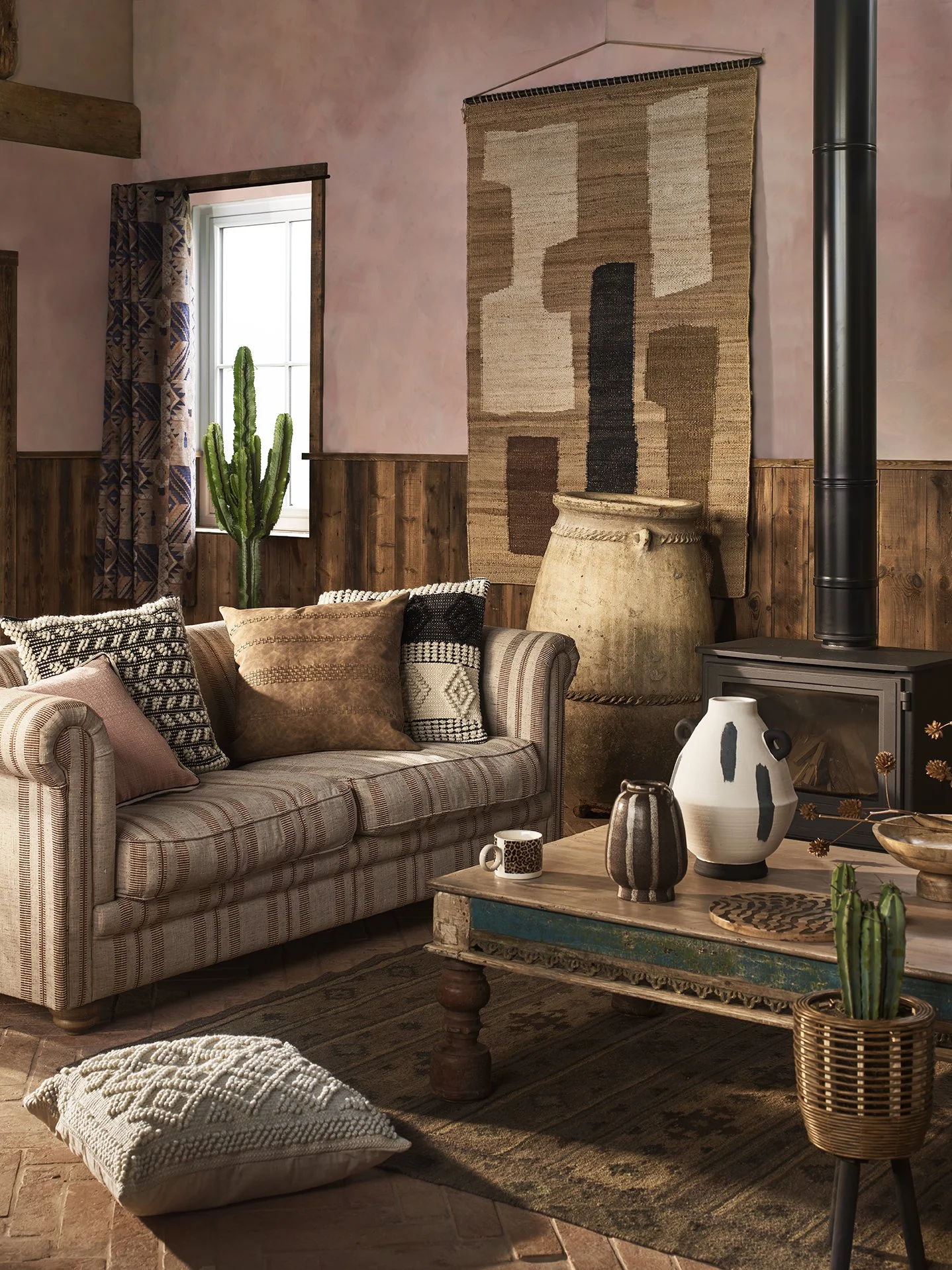 Cozy living room with a vintage striped sofa, decorative pillows, a rustic wooden coffee table with vases and mugs, a patterned pillow on the floor, a brick and wood-paneled wall with an abstract woven wall hanging, a window with patterned curtains, 