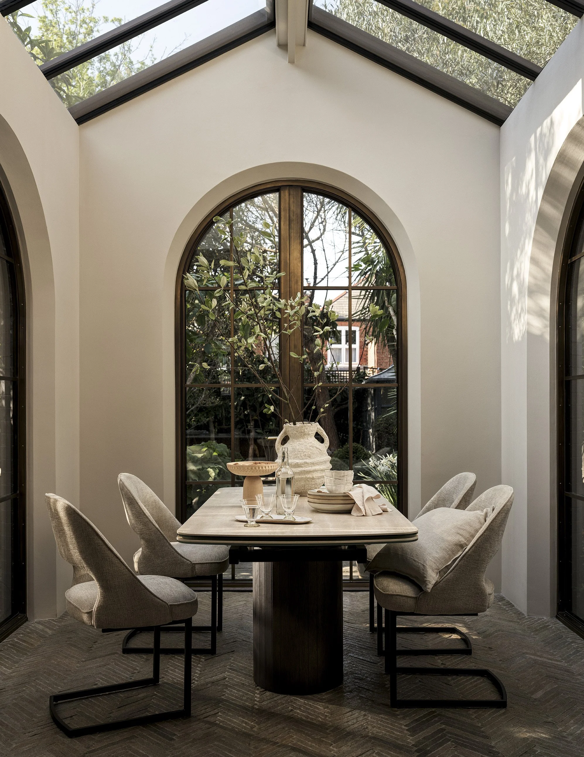 A dining nook with a wooden table, six cushioned chairs, and a large Arched window with greenery outside, inside a sunlit room.