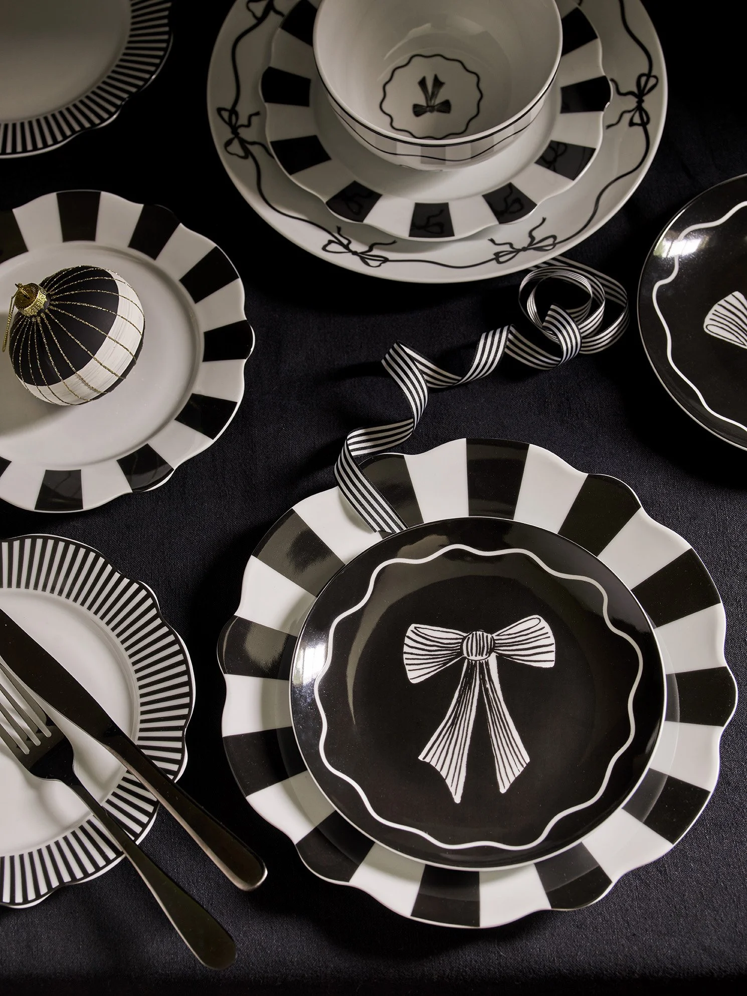 Black and white holiday-themed tableware with bow and stripe designs, including plates, cups, and a small ornament on a dark tablecloth.