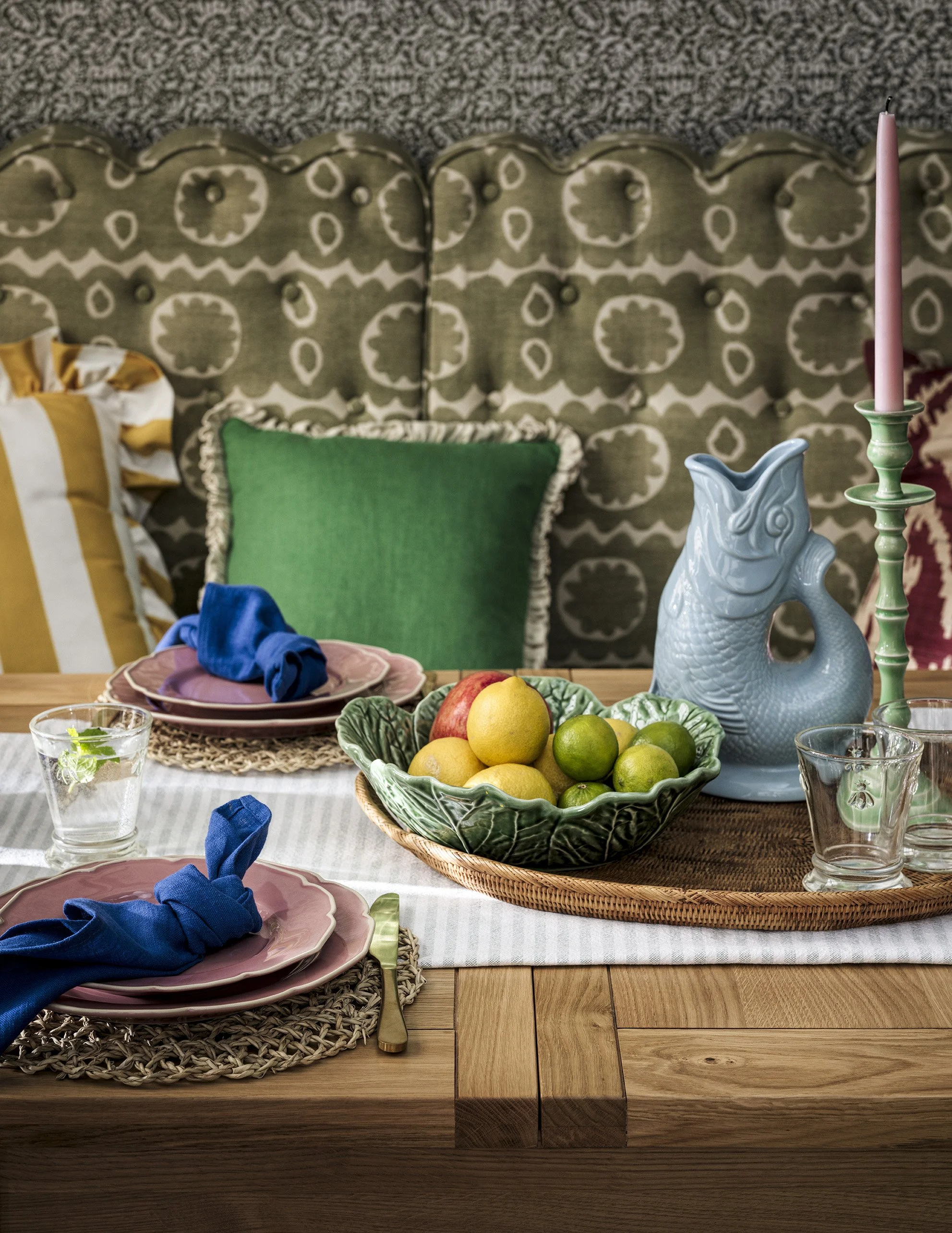 A dining table set with pink plates, blue cloth napkins, glasses of water, a bowl of lemons and limes, a decorative fish-shaped pitcher, a green candlestick with a pink candle, and colorful pillows in the background.