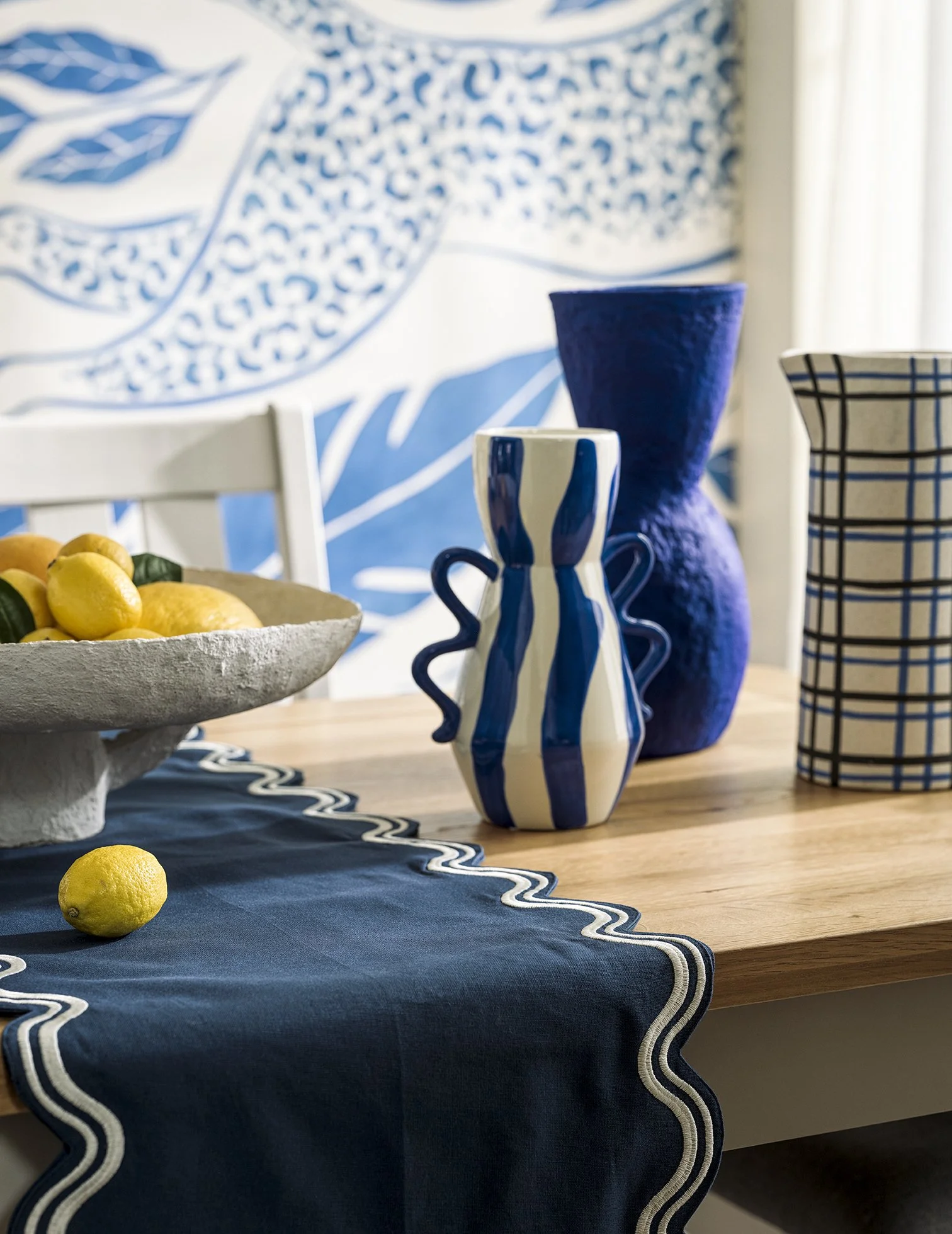 Decorative table setting with blue and white vases, a bowl of lemons, and a blue table runner, with a blue and white patterned wall in background.