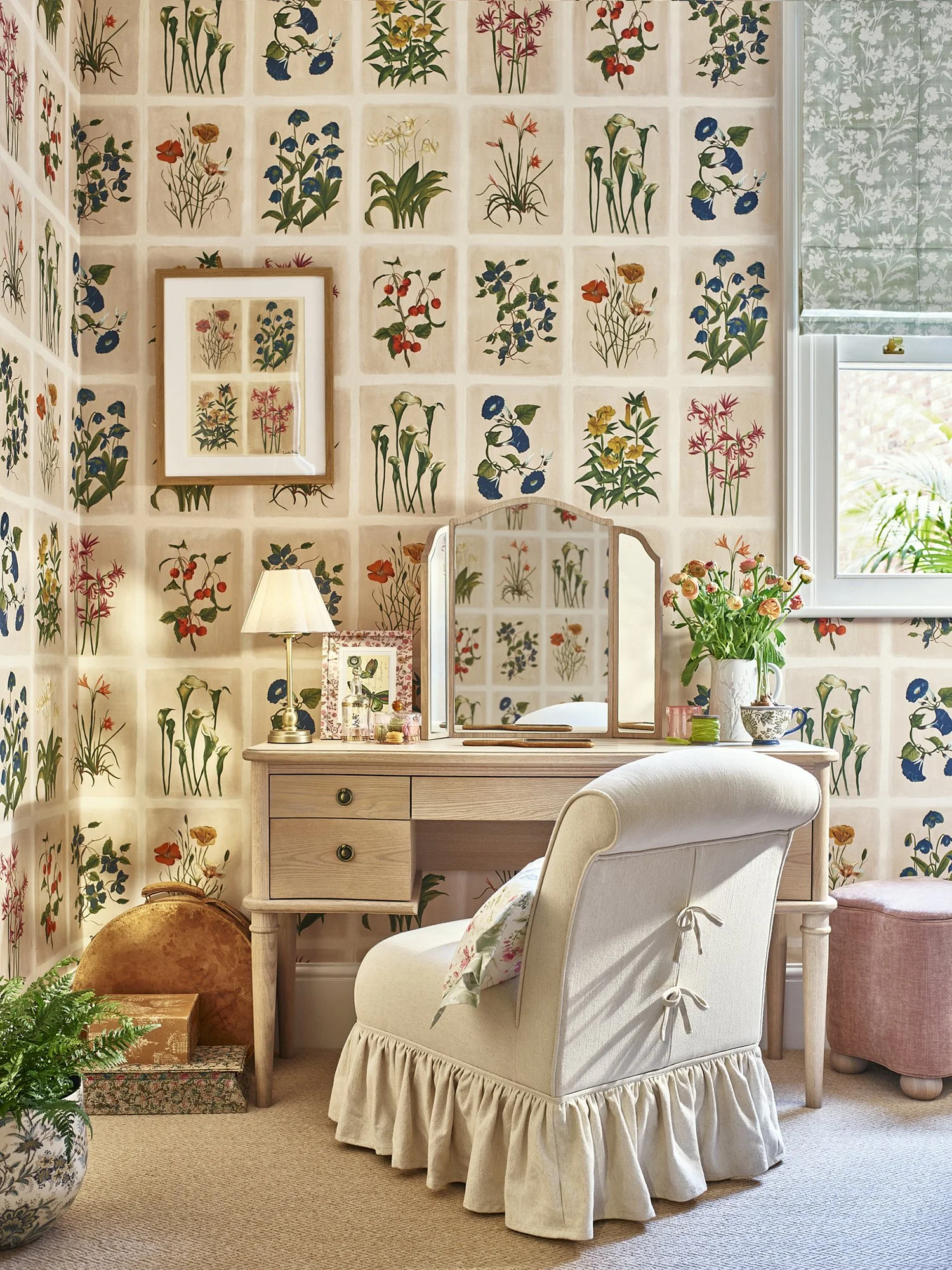 A cozy room decorated with floral wallpaper, a white desk with a mirror, a framed floral print, a table lamp, a vase of pink and orange flowers, and a white ruffled armchair.