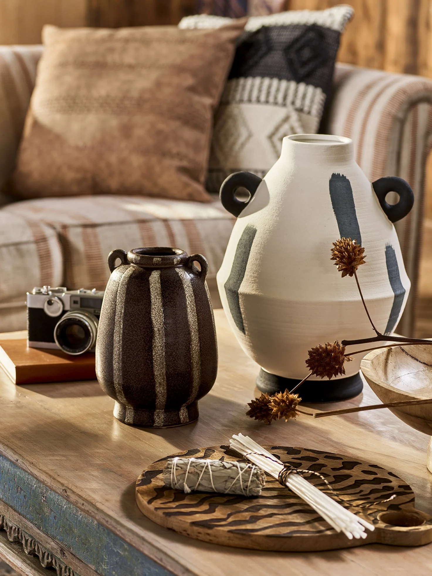Decorative beige and black vases, dried flowers, a bundle of white sticks, a small camera, and a wooden tray on a rustic wooden table in a cozy living room with striped and cushion pillows.