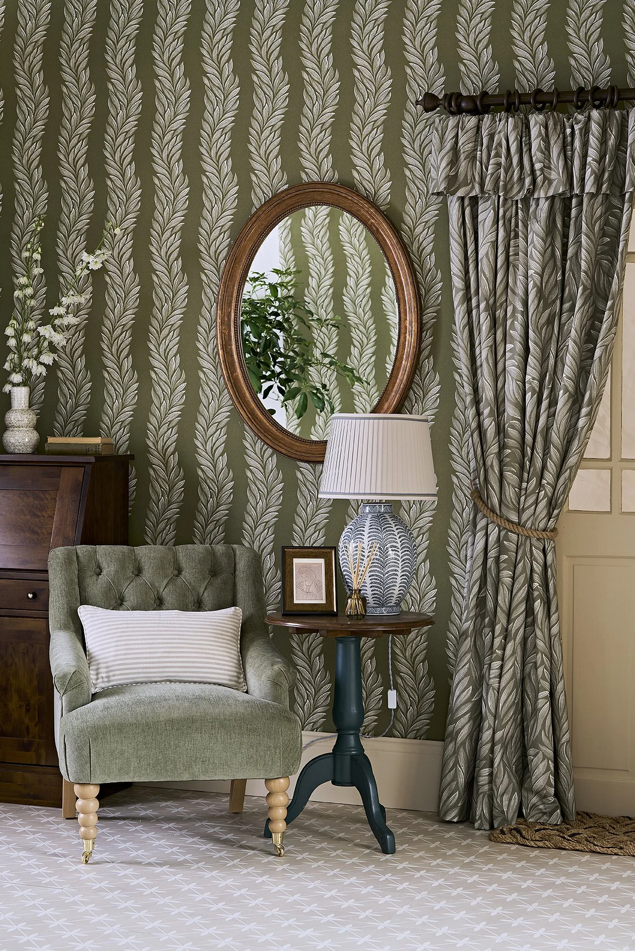 A cozy living room corner with a green tufted armchair, a side table with a peacocks pattern lamp, framed artwork, and a reed diffuser. The wall has a leafy green wallpaper, a round mirror, and a patterned curtain.