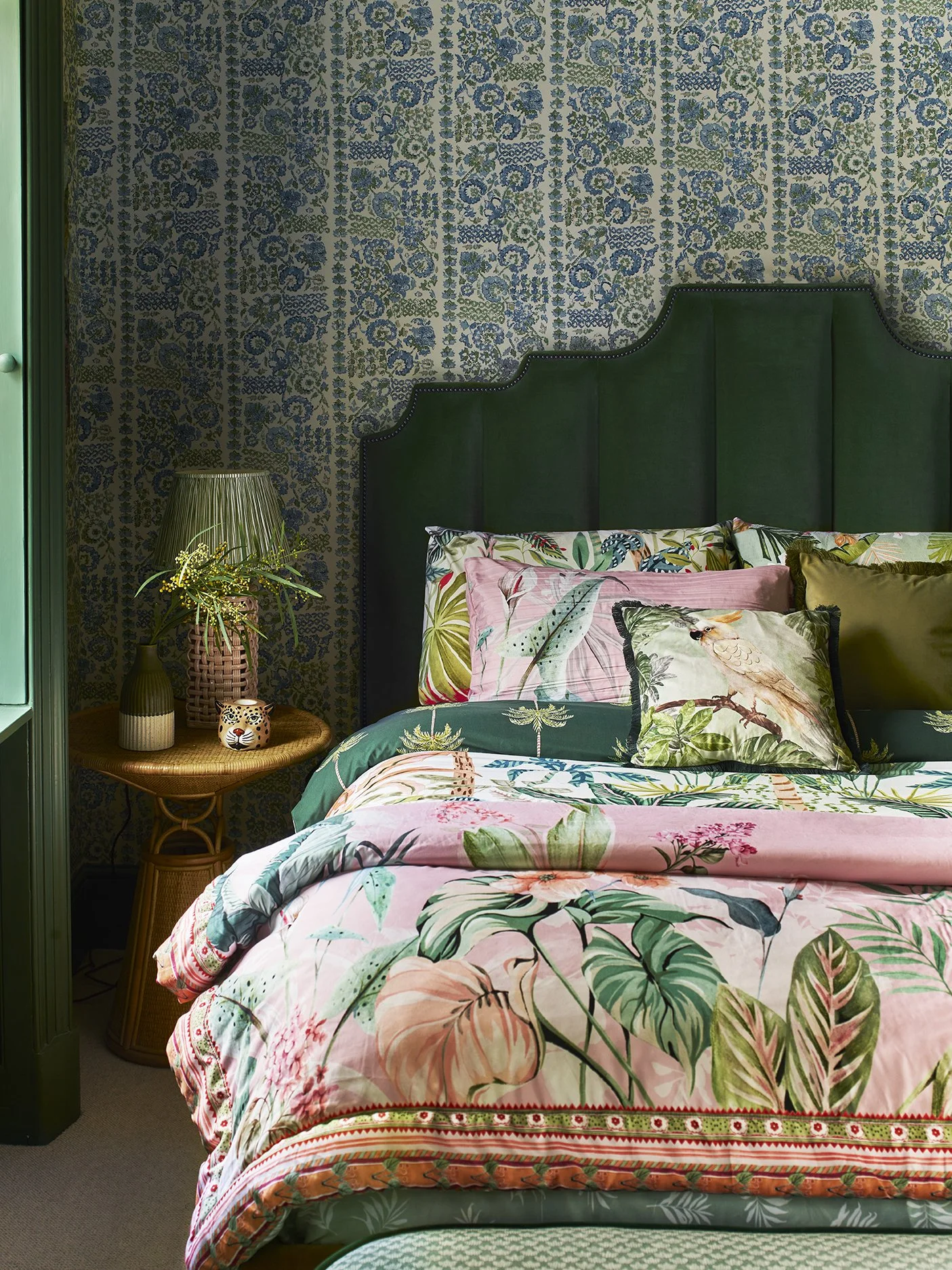 A bedroom with tropical-themed bedding featuring large floral and leaf patterns, a dark green upholstered headboard, a wicker nightstand with a plant and decorative items, and a patterned wallpaper with blue and green floral motifs.