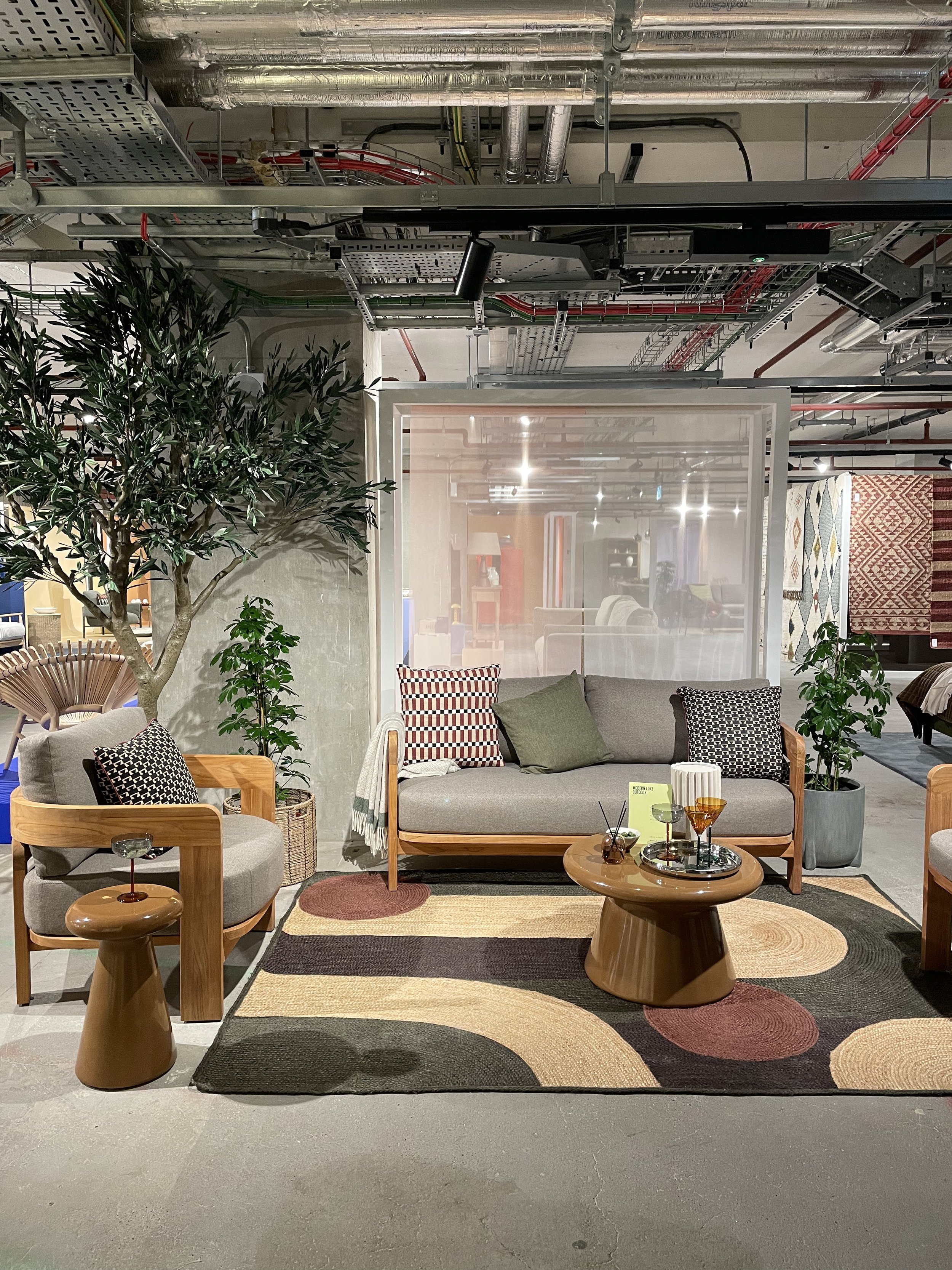 A stylish indoor furniture display featuring a gray sofa with wooden armrests and patterned throw pillows, two matching armchairs, a round wooden coffee table with drinks, a patterned area rug, potted plants, and other furniture pieces visible in the