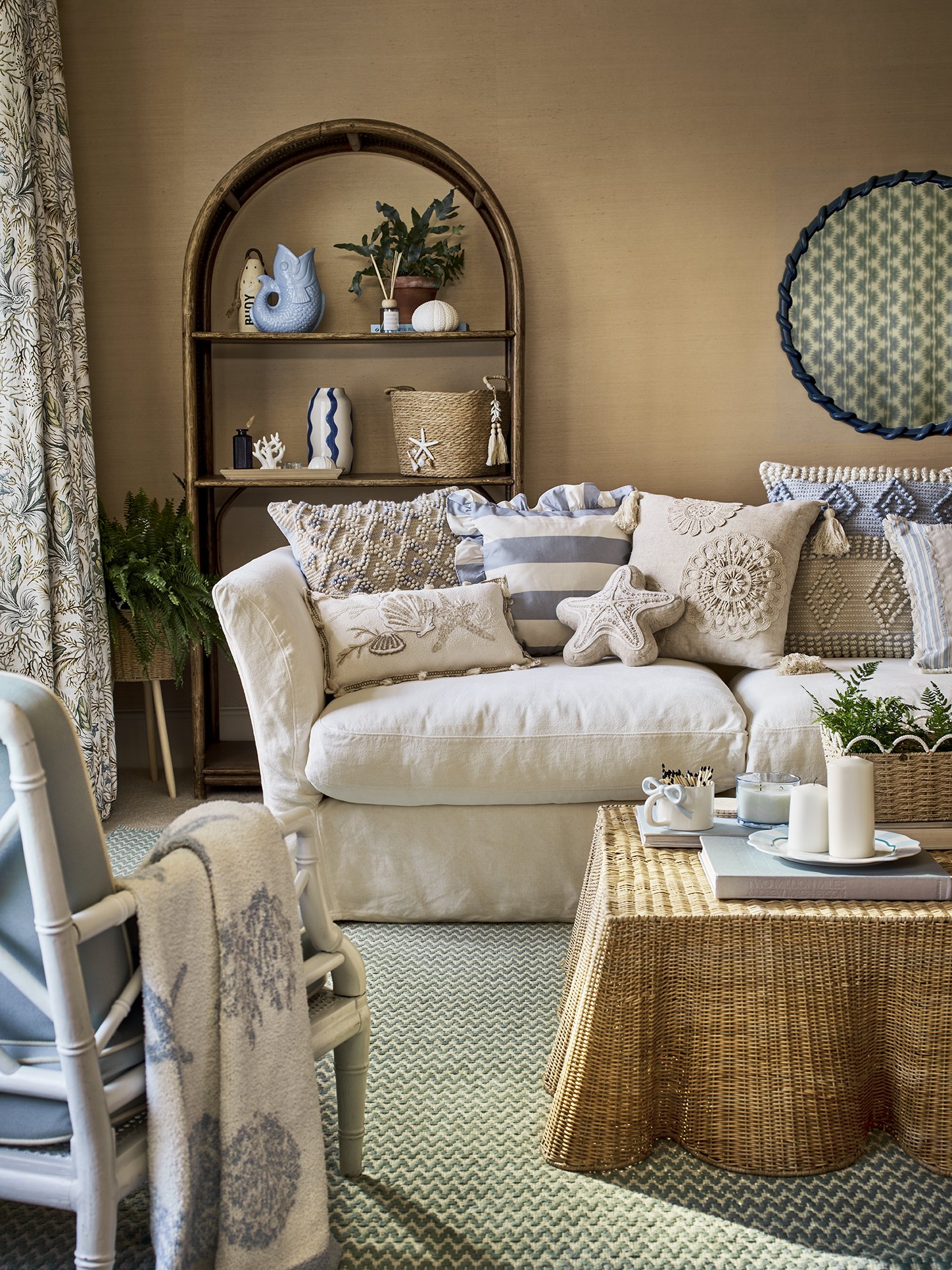 Cozy living room with a white sofa decorated with blue and beige pillows, a wicker coffee table with candles and decor, and a wooden shelf with decorative vases, plant, and starfish, all with coastal theme.