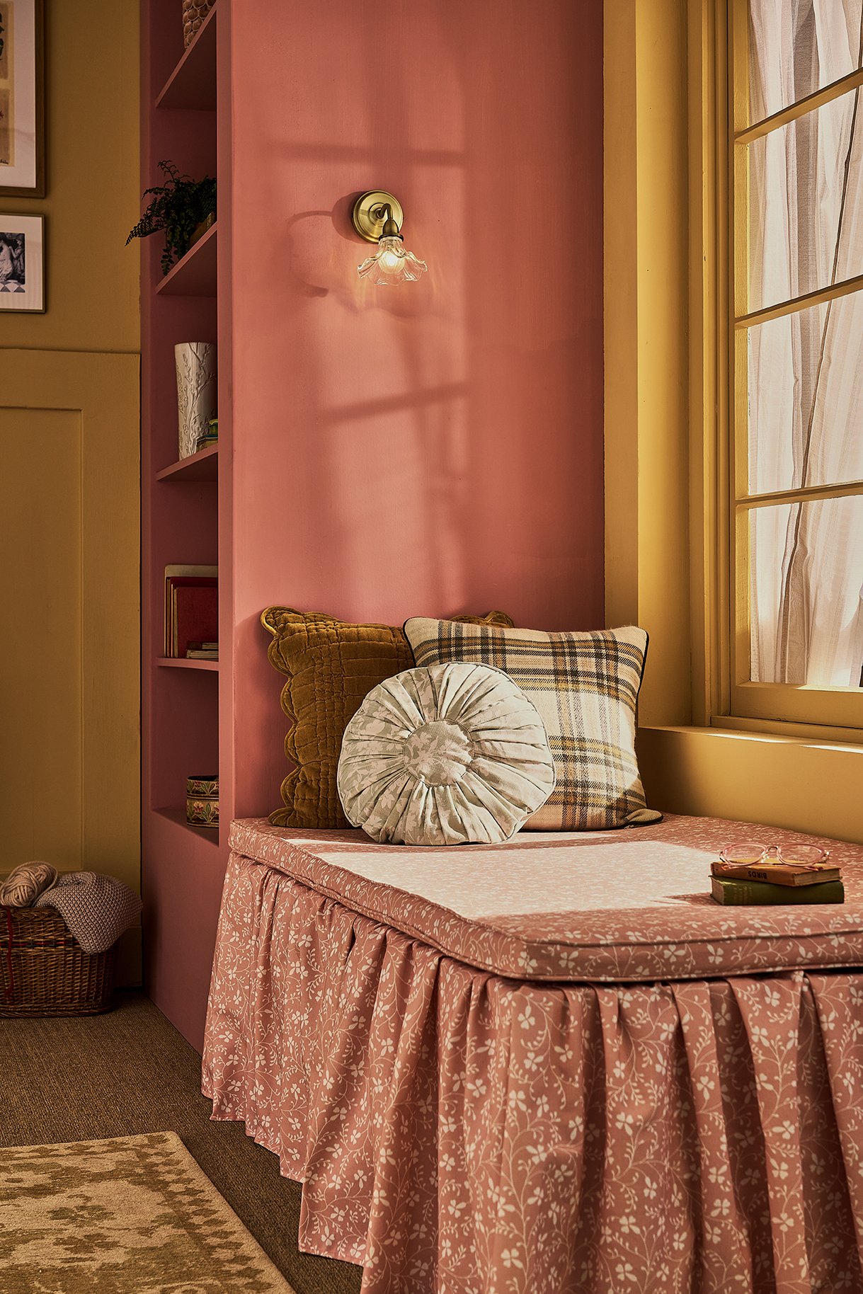 A cozy window seat with pink floral upholstery, decorated with patterned and velvet throw pillows, next to a window with wooden framing, in a room with pink and yellow walls and a built-in bookshelf.