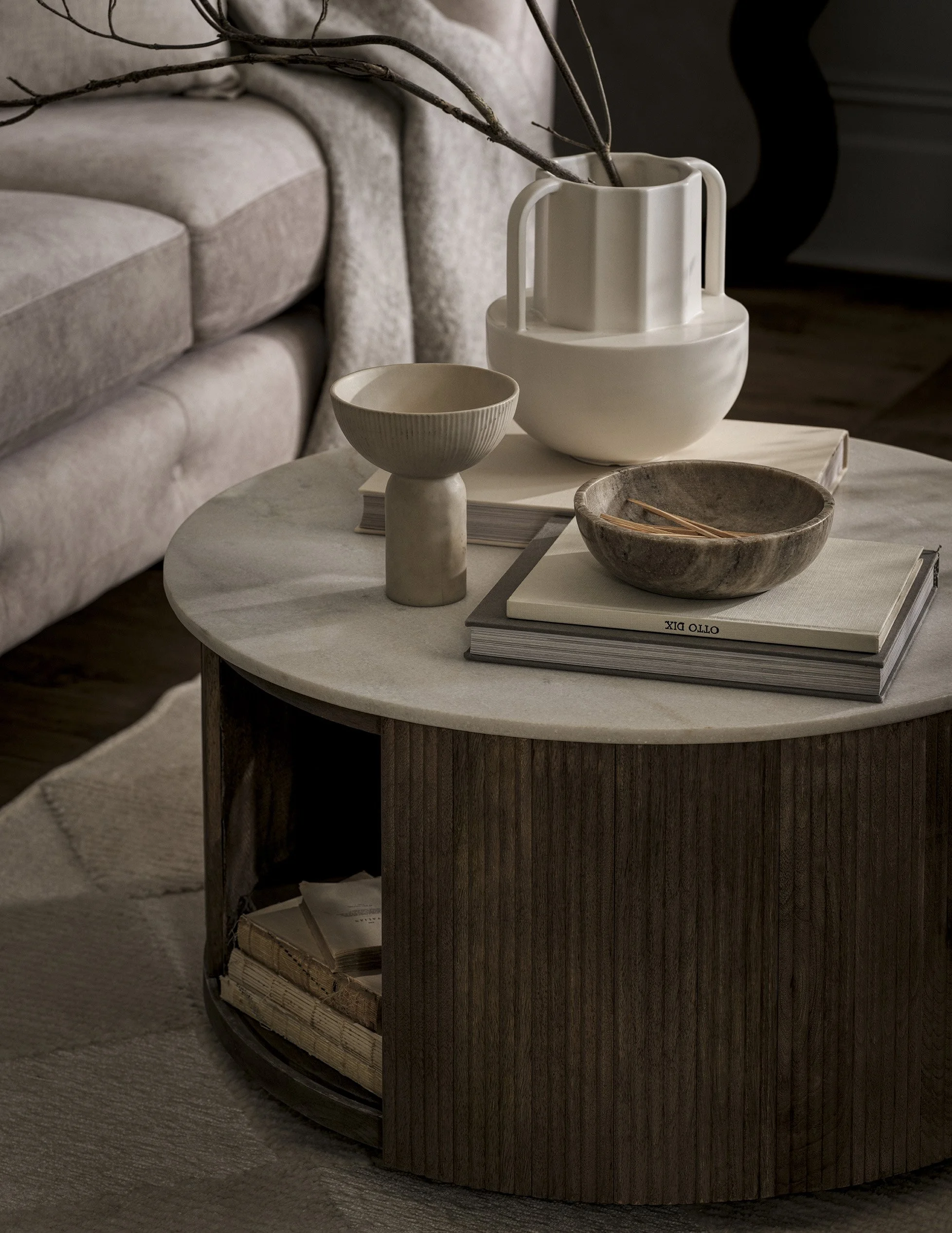 Round wooden coffee table with a white marble top, decorated with books, a ceramic bowl, a stone bowl with chopsticks, and a white ceramic jug with branches. A sofa with light-colored fabric is in the background.
