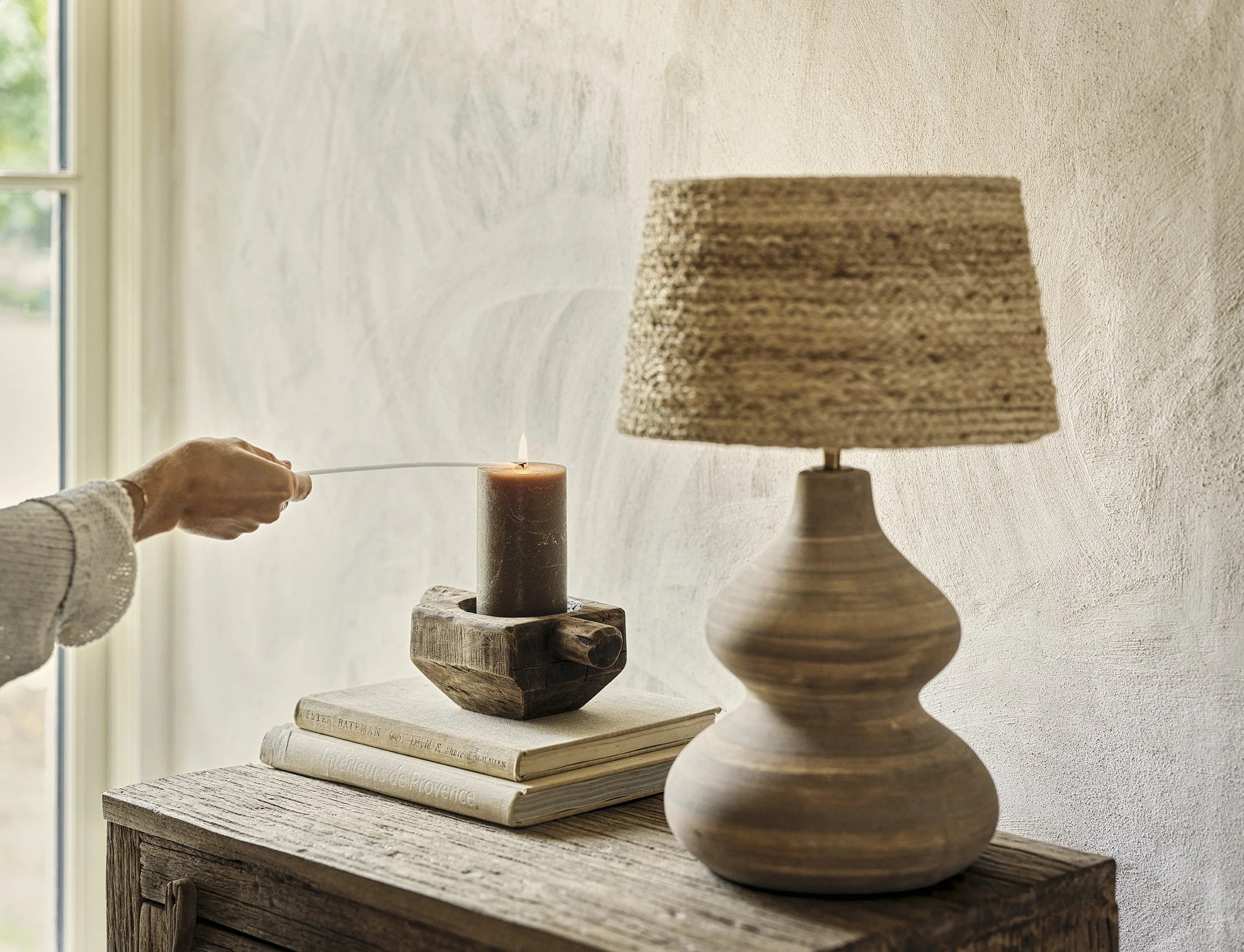 Person lighting a candle on a wooden table near a window, with a lamp and stacked books nearby.