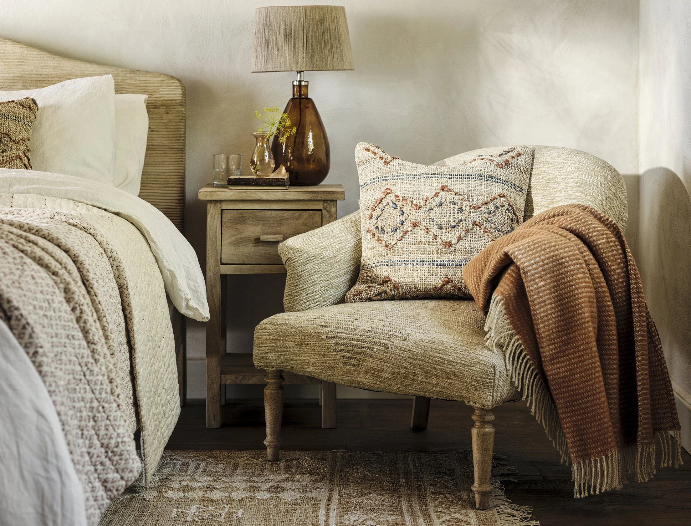 A cozy bedroom corner with a wooden headboard bed, a side table with a lamp and decorative items, and an upholstered armchair with a patterned pillow and a rust-colored throw blanket.
