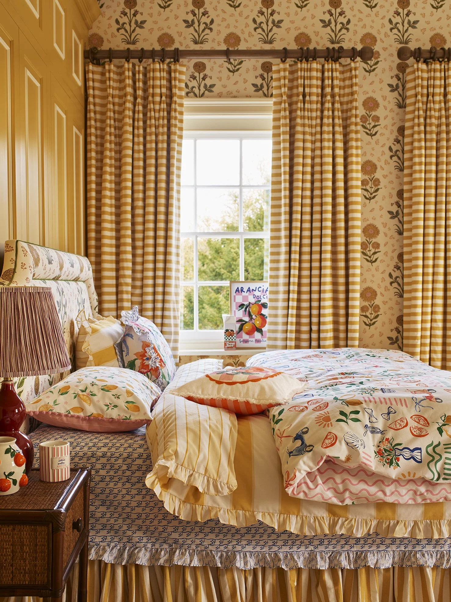 A cozy bedroom with floral wallpaper, yellow and white striped curtains, a bed with colorful patterned pillows, a wooden side table with a striped candle, and a large window showing green trees outside.