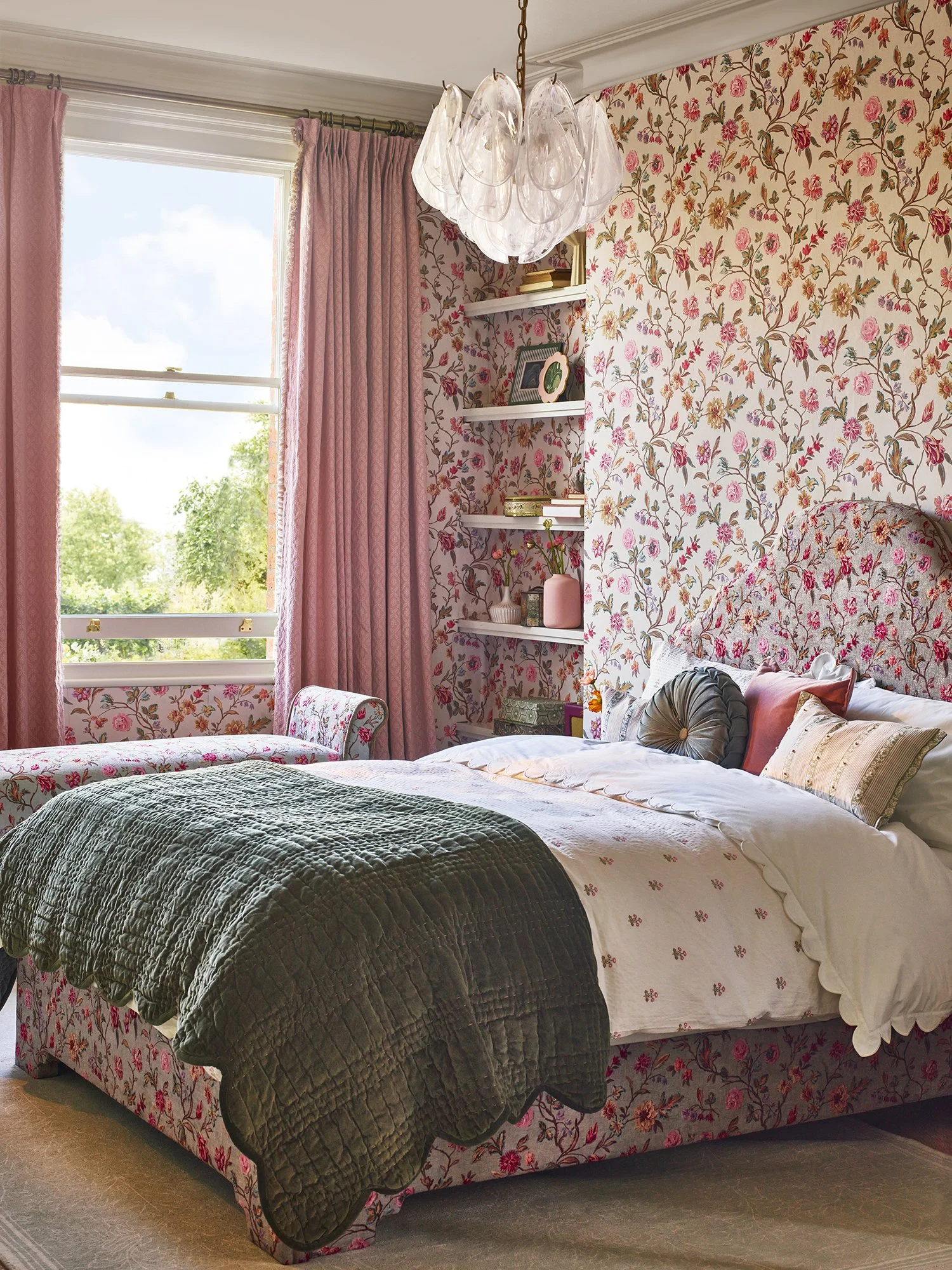 A bedroom with floral wallpaper, pink curtains, and a bed with white and pink bedding. There is a chandelier hanging from the ceiling, a window with green trees outside, and shelves with decorative items.
