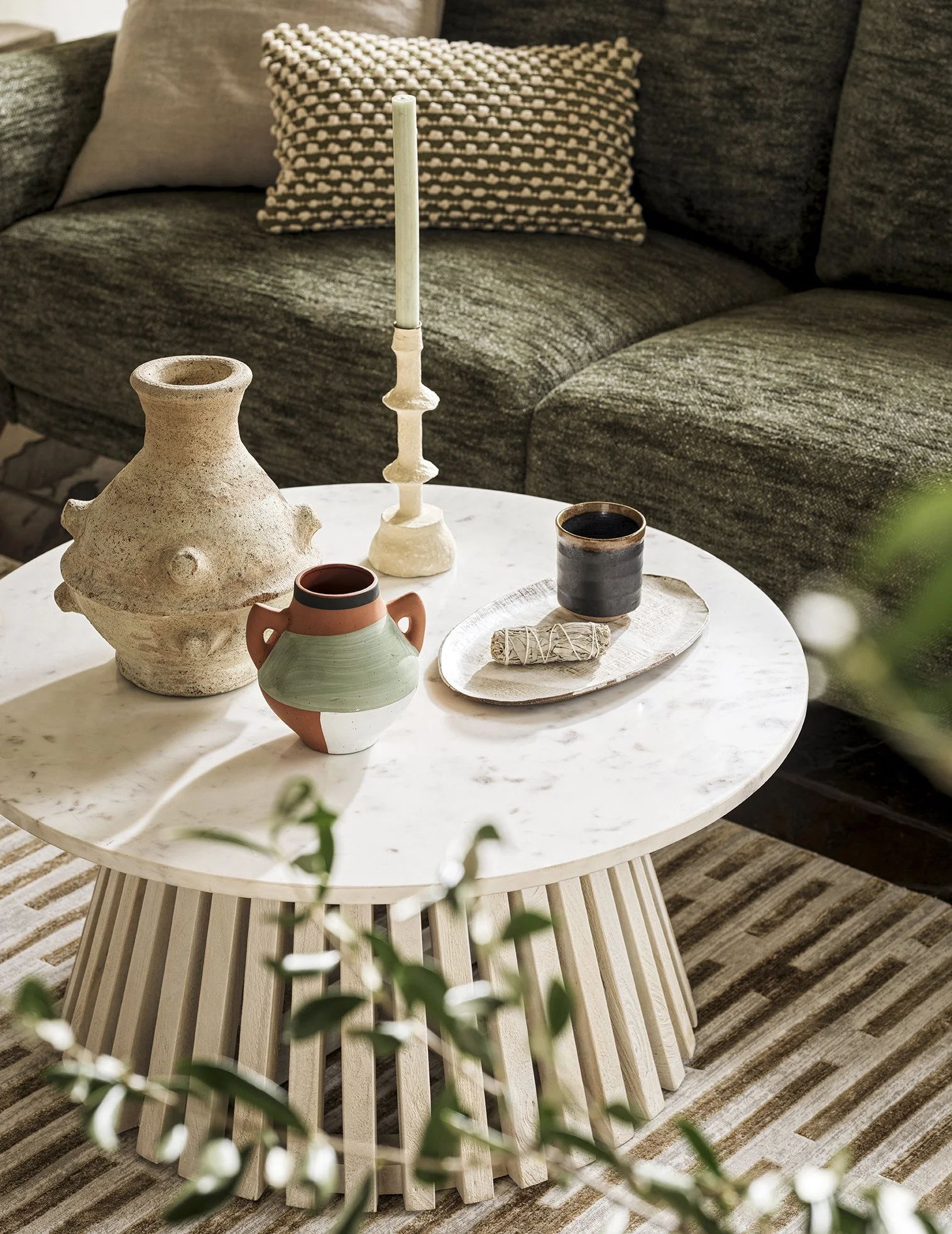 Decorative items on a white marble coffee table in front of a dark green sofa with a patterned pillow. Items include a large textured ceramic vase, a small green and white vase, a beige candlestick with a tall white candle, a black ceramic cup, and a