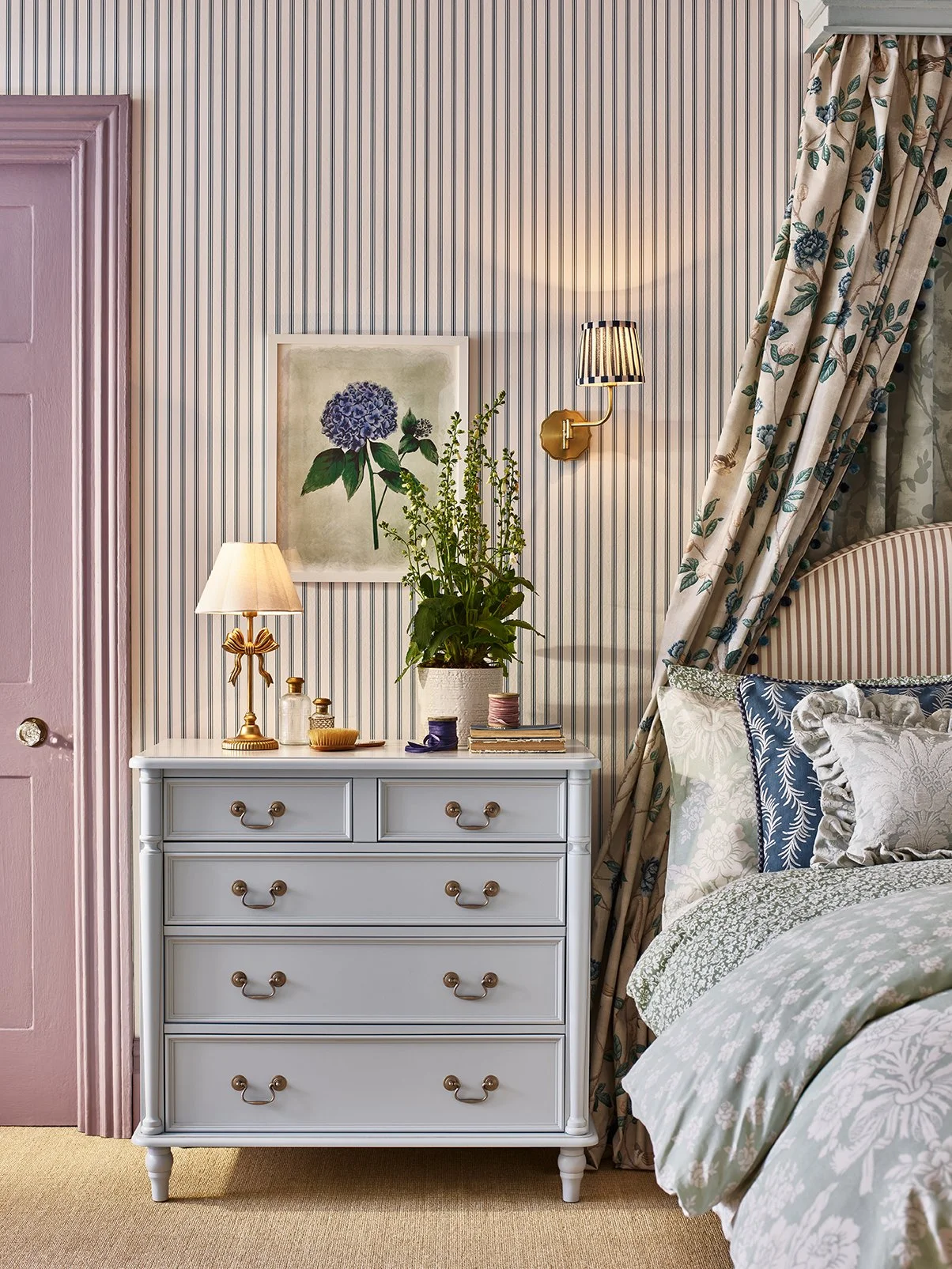 A bedroom corner with a white dresser, a potted plant, a table lamp, and framed artwork of a blue hydrangea. A bed with patterned bedding and floral curtains is partially visible.