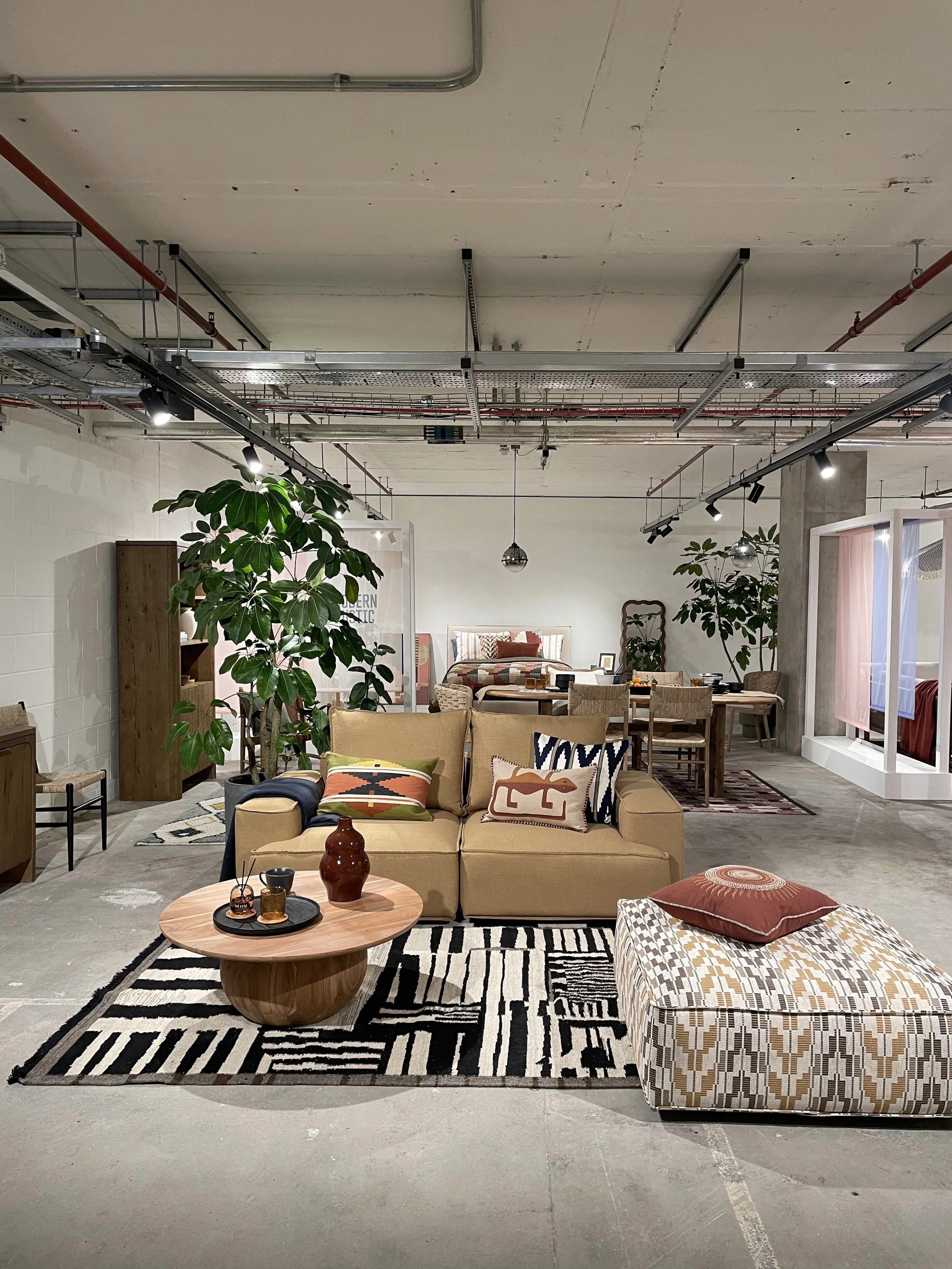 Modern furniture arrangement in a spacious room with a beige sofa, patterned pillows, a round wooden coffee table with decorative items, a patterned ottoman, large green plants, and a dining area in the background.