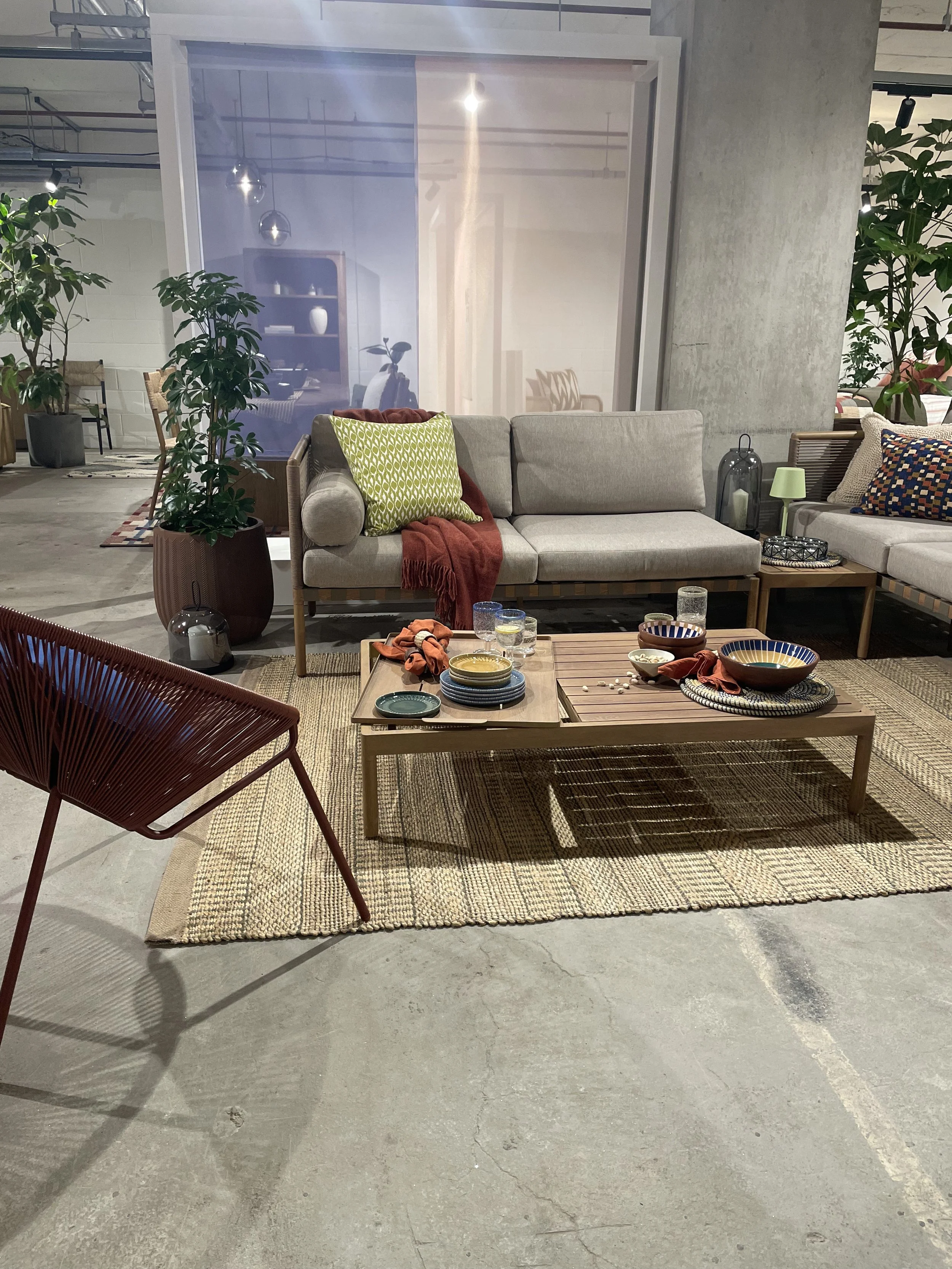 Living room with beige couch, red and green pillows, wooden coffee table with bowls and glasses, red chair, patterned rug, potted plant, decorative lamps, and another seating area.