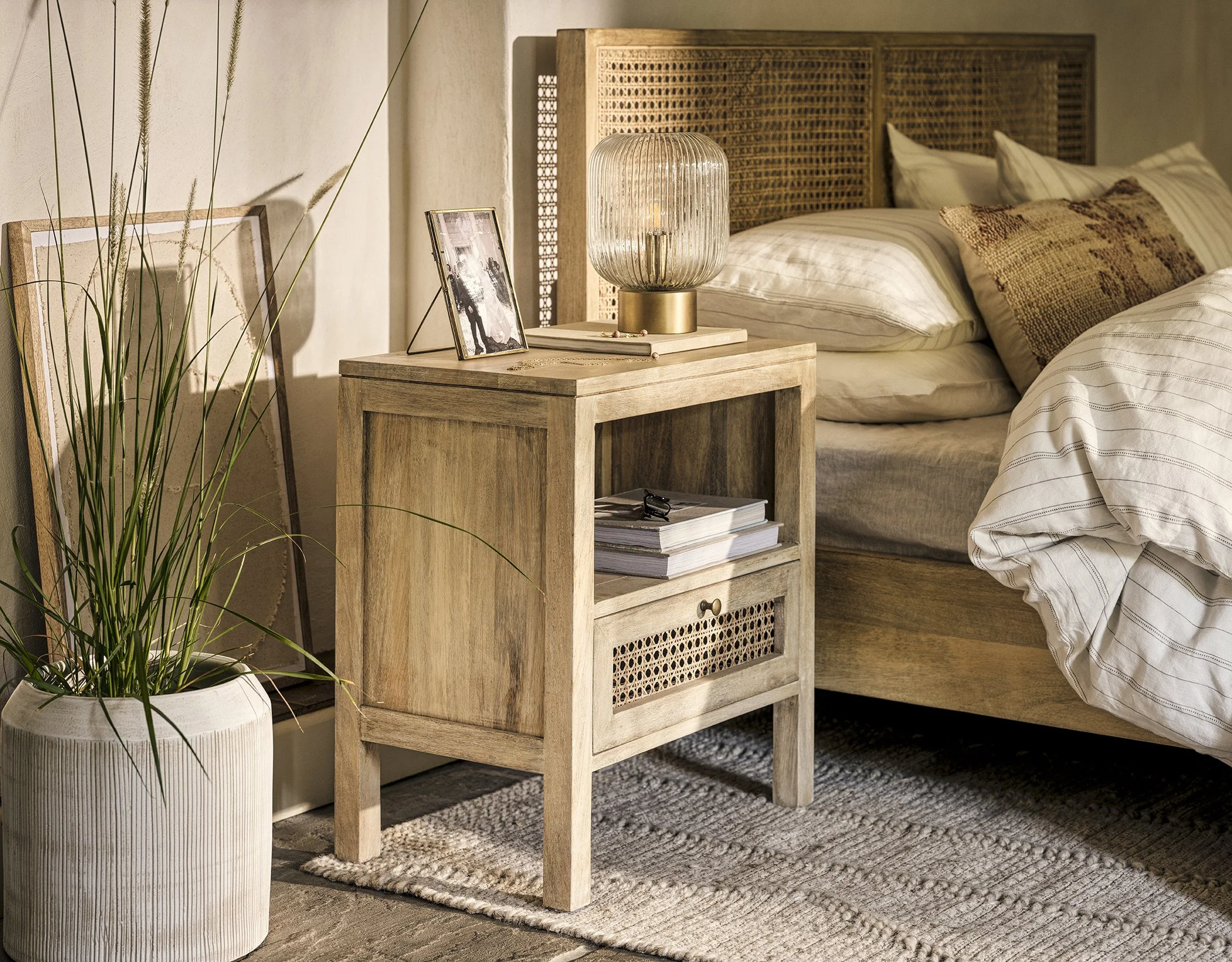 A cozy bedroom featuring a wooden nightstand next to a bed with striped bedding, a lamp, framed photo, and a book on the nightstand, with potted plant and framed artwork nearby.