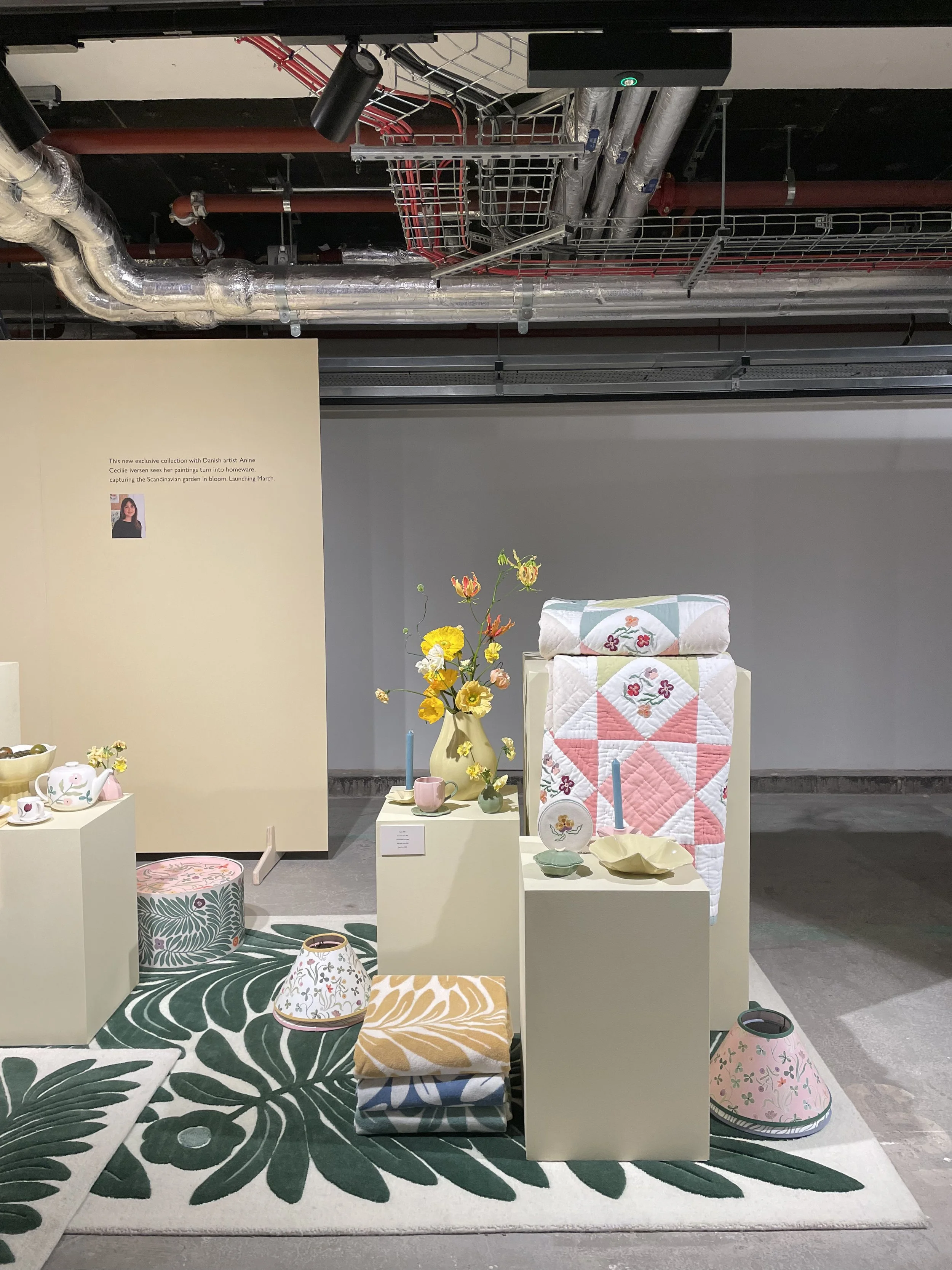 Display of various home decor and textile items, including patterned blankets, lamps, a vase with flowers, candles, dishes, and cushions, arranged on pedestals and a carpet with a large green leaf design.