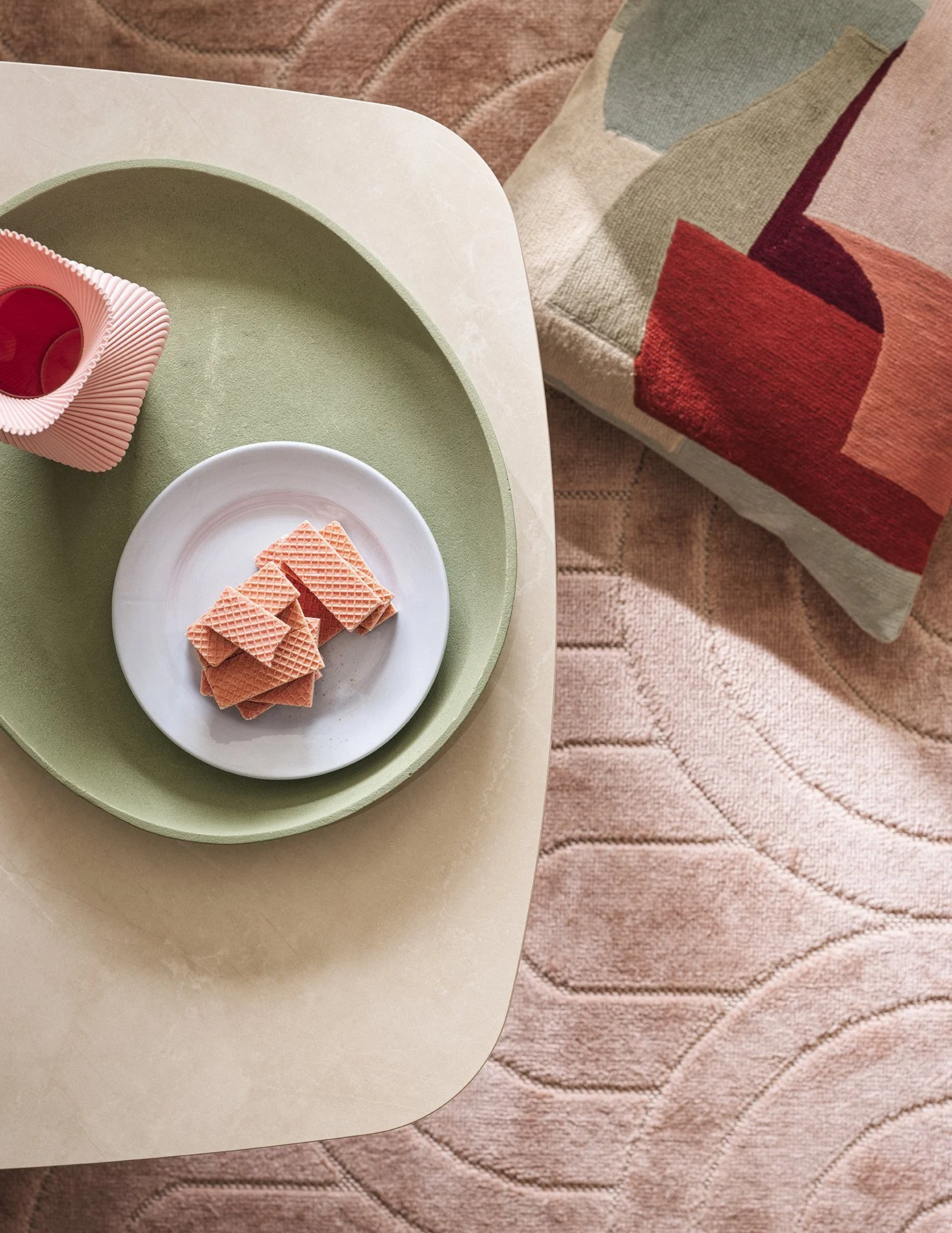 A top-down view of a beige table with a green placemat, a white plate of pink waffle biscuits, and a pink textured cup, with a pink patterned cushion on a pink carpet in the background.