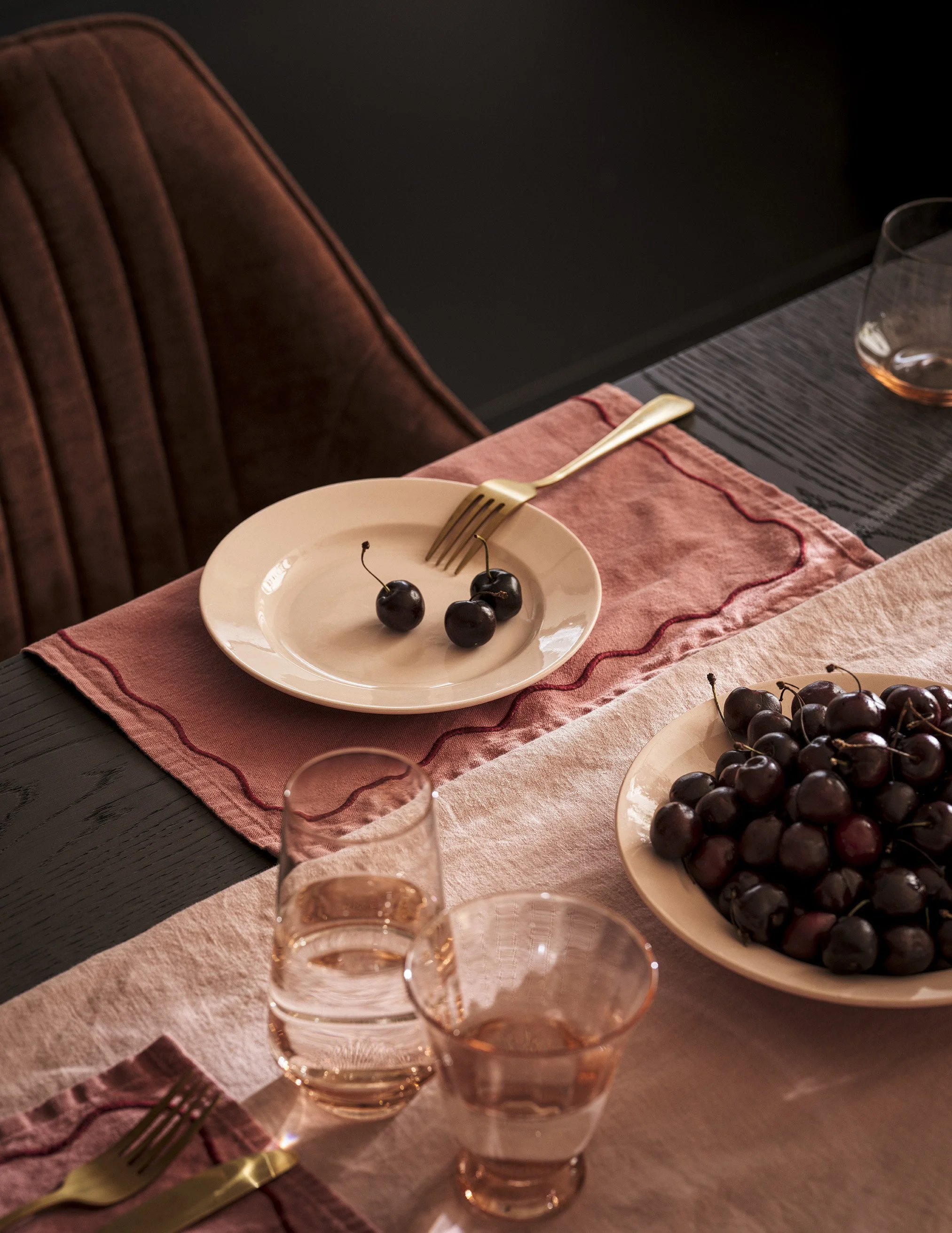 A dining table set with a pink table runner, gold utensils, a plate with three cherries, a large bowl of cherries, and two glasses of rosé wine in a dimly lit room.