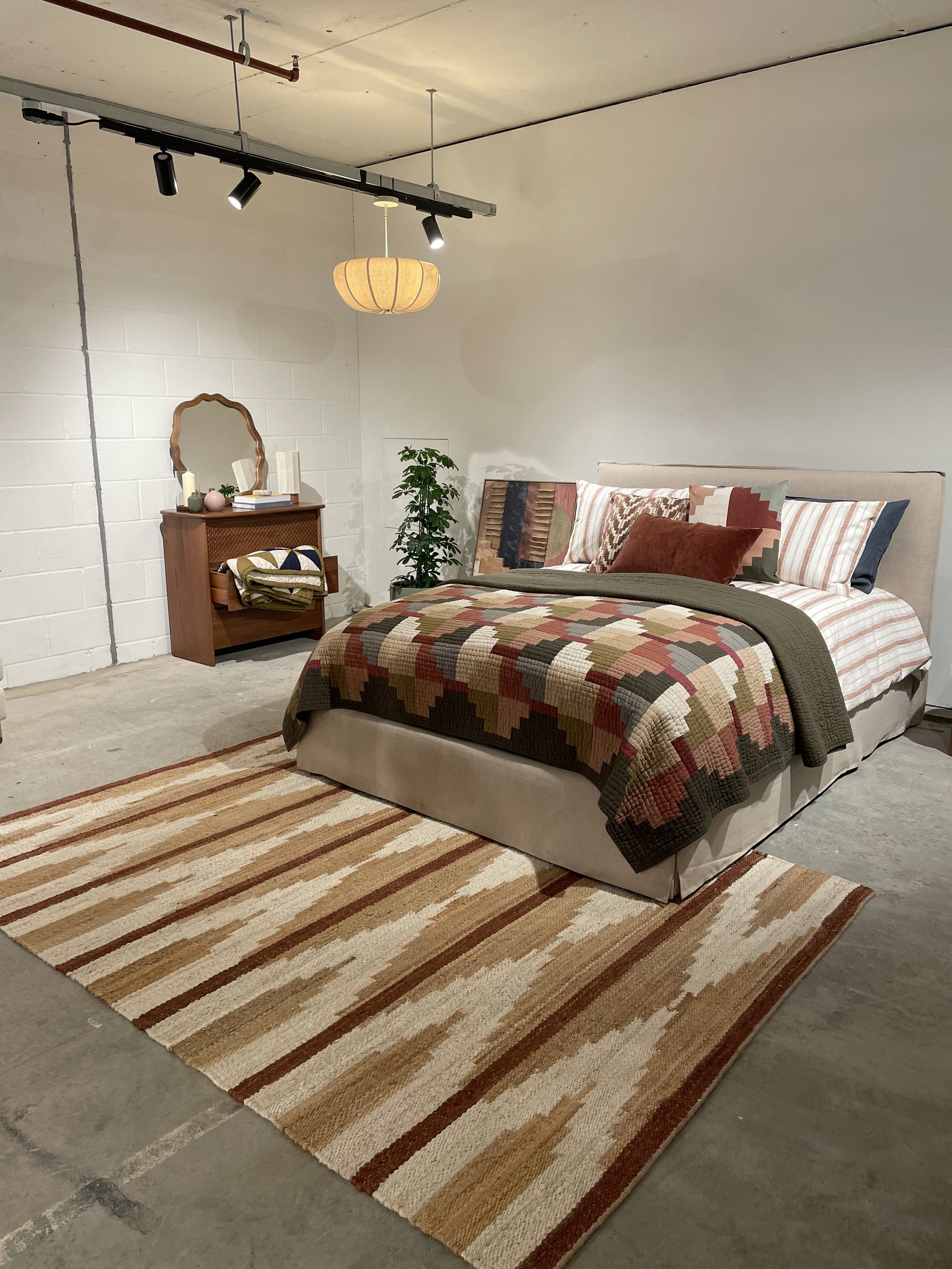 A bedroom with a beige bed, patterned quilt, pillows, a wooden dresser, mirror, potted plant, hanging light, and striped area rug.