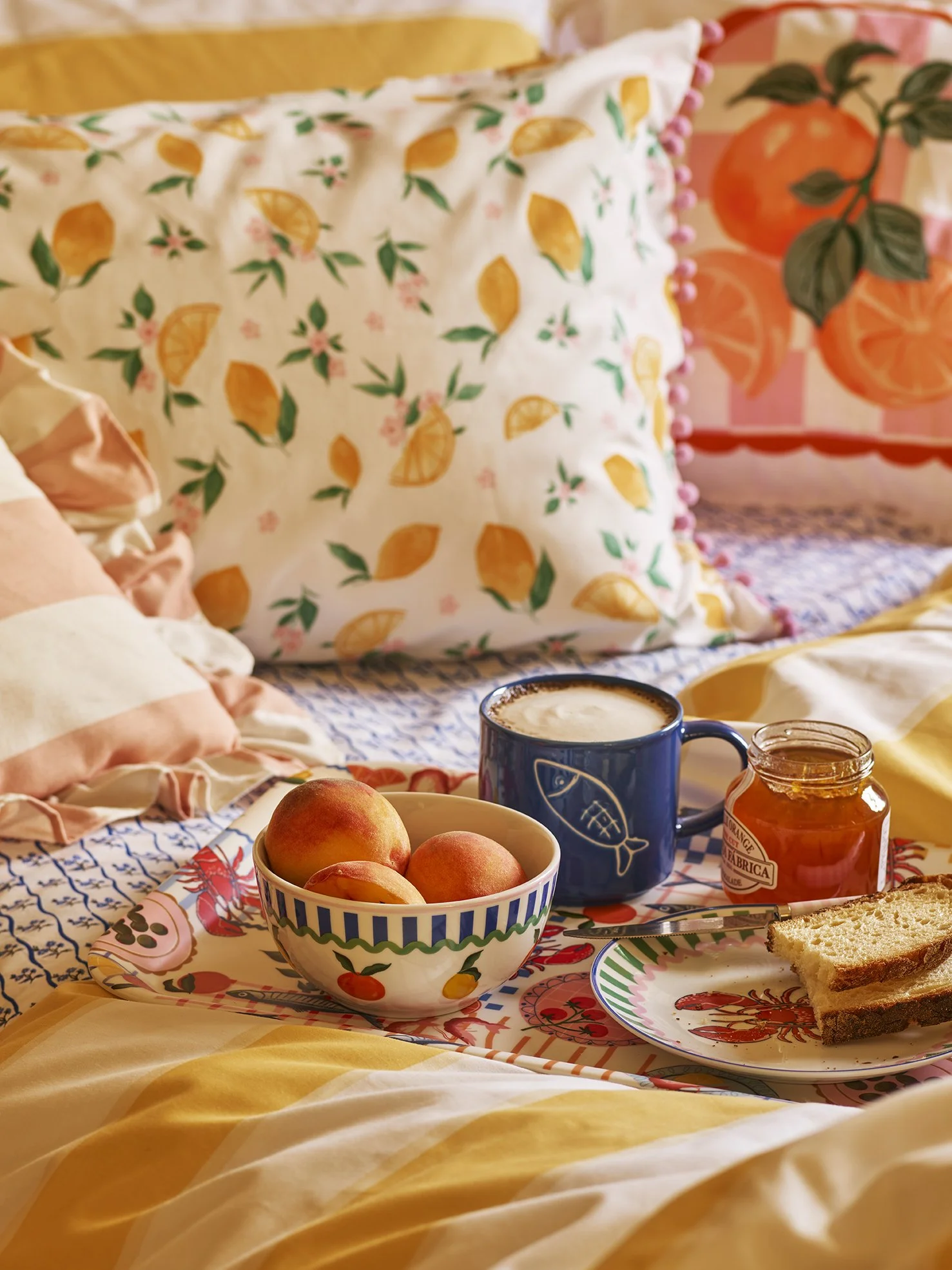 Breakfast in bed with peaches, a cup of coffee, toast, and jam on decorative colorful plates, with floral and citrus-themed pillows in the background.