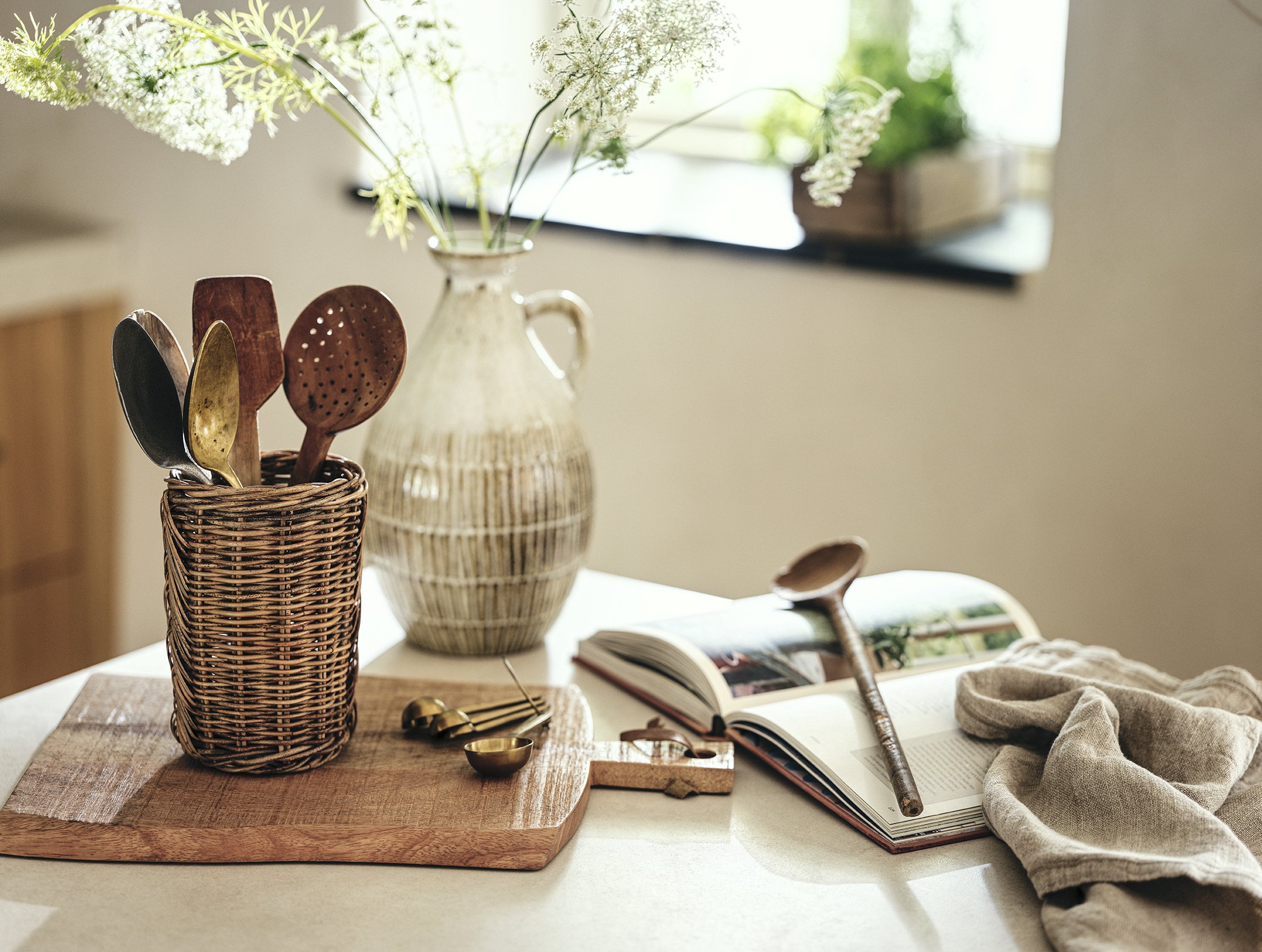Kitchen table with a wicker utensil holder containing metal and wooden spoons, a ceramic vase with white flowers, an open magazine, a scoop, and a beige cloth.