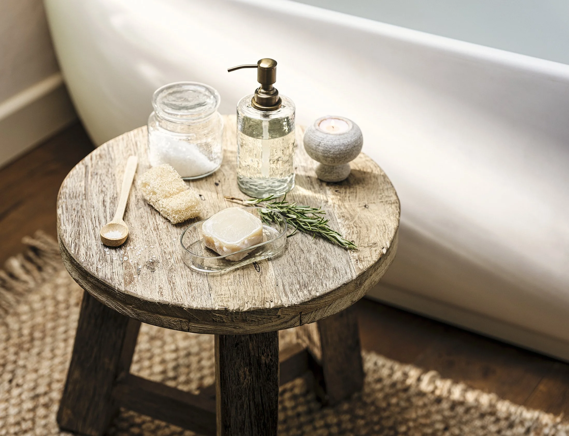 A rustic round wooden stool with spa essentials, including a soap bar, a small dish, a wooden spoon, a jar of salt, a soap dispenser, a candle holder, and some sprigs of rosemary, near a white bathtub and a woven rug.