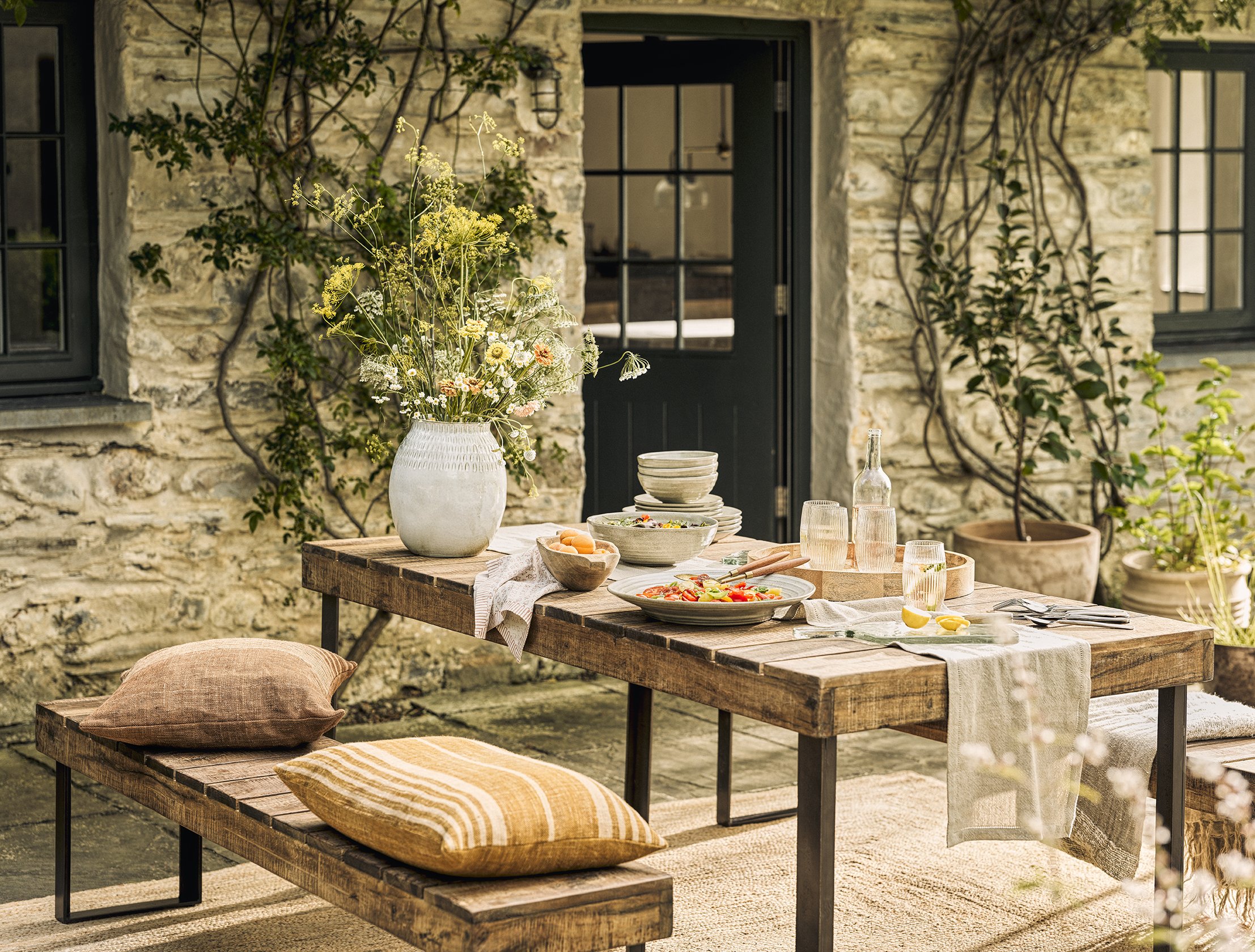 Outdoor dining table set with plates, glasses, a large flower vase, and cushions on a wooden bench against a stone wall with windows and climbing plants.