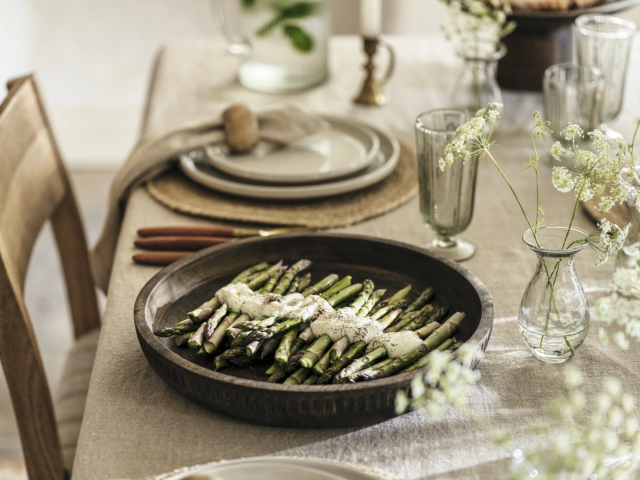 A dining table with a large black bowl of cooked asparagus topped with a creamy sauce, set with plates, glasses, and decorative flowers in a glass vase, in a cozy, rustic style dining room.